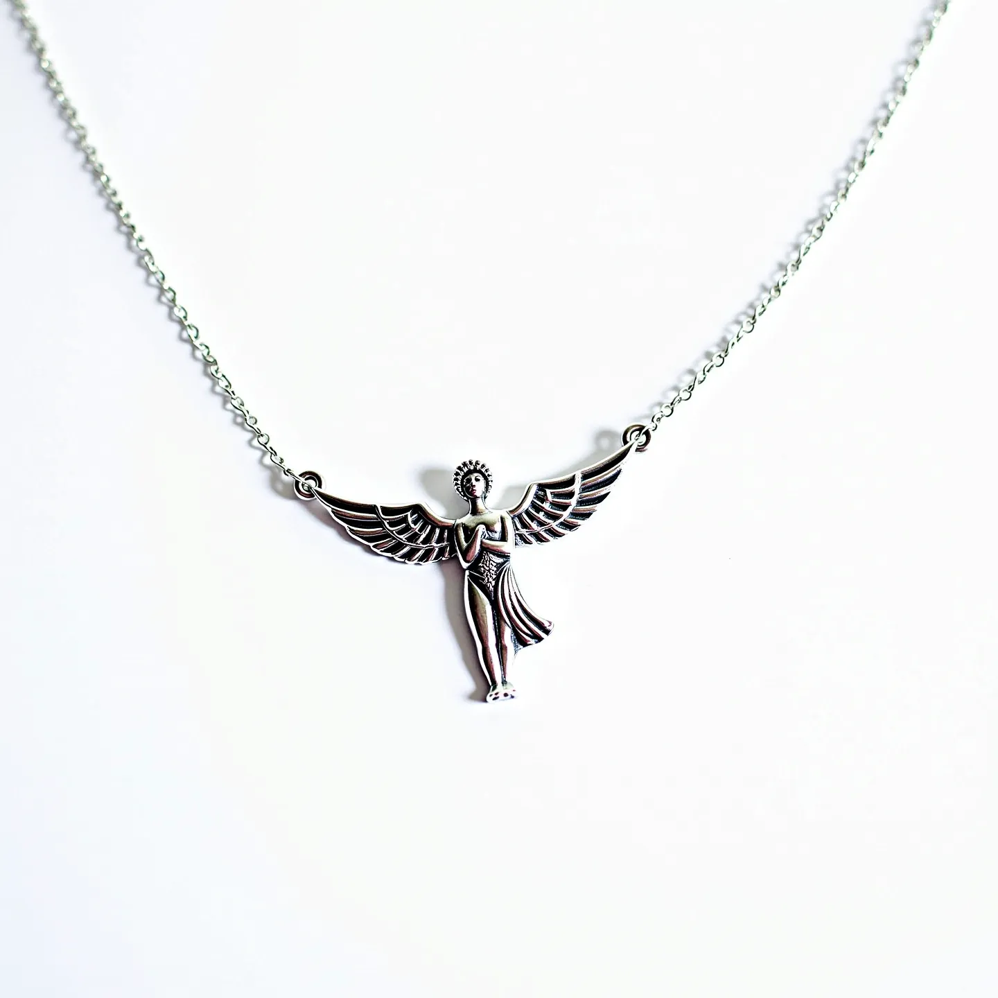 This angel necklace features a finely detailed angel pendant crafted from a silver-toned metal, displaying intricate wings and a flowing gown. The chain is composed of uniformly sized round links, contributing to a delicate and elegant appearance. The pendant is seamlessly integrated into the chain with metal rings attached to each wing, ensuring a balanced presentation. The necklace likely includes a standard clasp mechanism at the back, typical for securing necklaces of this style. The overall design emphasizes grace and serenity, characteristic of angelic symbolism.