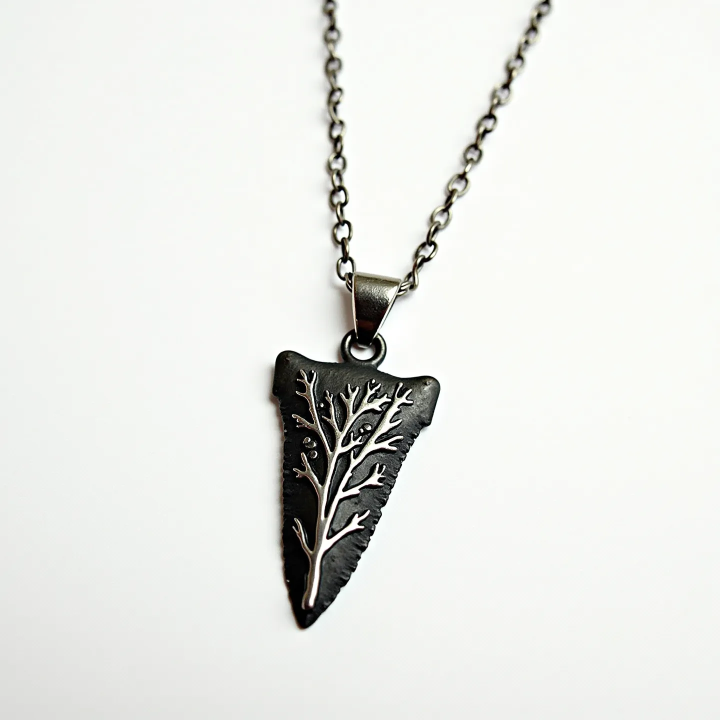 This arrowhead necklace features a pendant crafted from a dark metal, possibly stainless steel or oxidized silver, with a matte finish that highlights an intricate, embossed tree or branch design on the surface. The necklace includes a simple chain composed of uniformly sized links, matching the dark tone of the pendant. The pendant is secured to the chain with a sturdy metal bail, which is attached through a loop at the top of the arrowhead. The necklace is closed with a standard clasp, ensuring ease of wear and security.