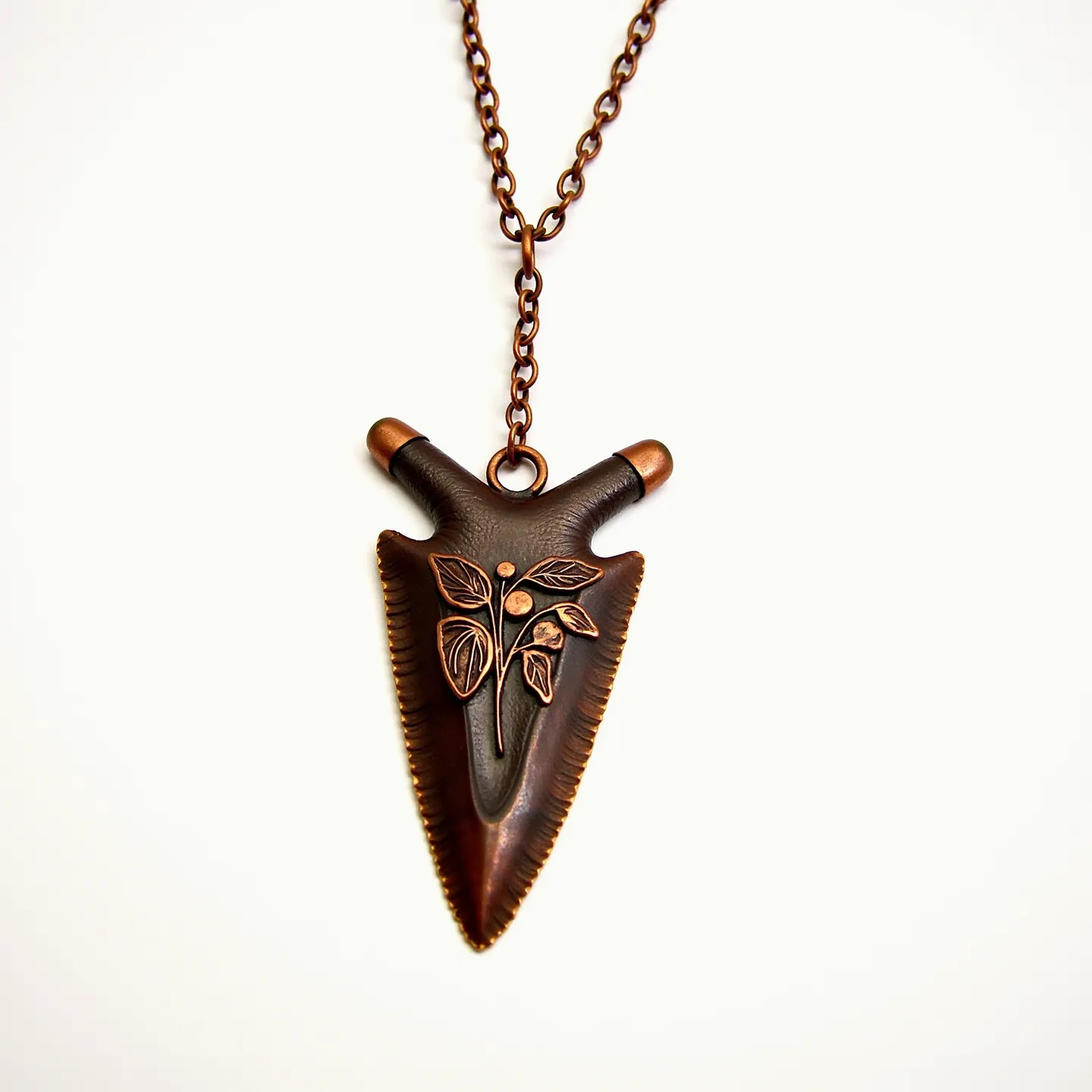 This arrowhead necklace features a pendant crafted from a metal that appears to have a bronze or copper finish, highlighted by an intricate engraved leaf design on its surface. The pendant is suspended from a matching metal chain, suggesting a cohesive and rustic aesthetic. The chain connects to the pendant through a simple loop attachment at the top of the arrowhead, ensuring a secure hold. There are no visible gems or stones set in the pendant, allowing the metalwork to be the primary focus. The closure of the necklace is not visible, but it likely utilizes a standard clasp consistent with the material and style of the piece.