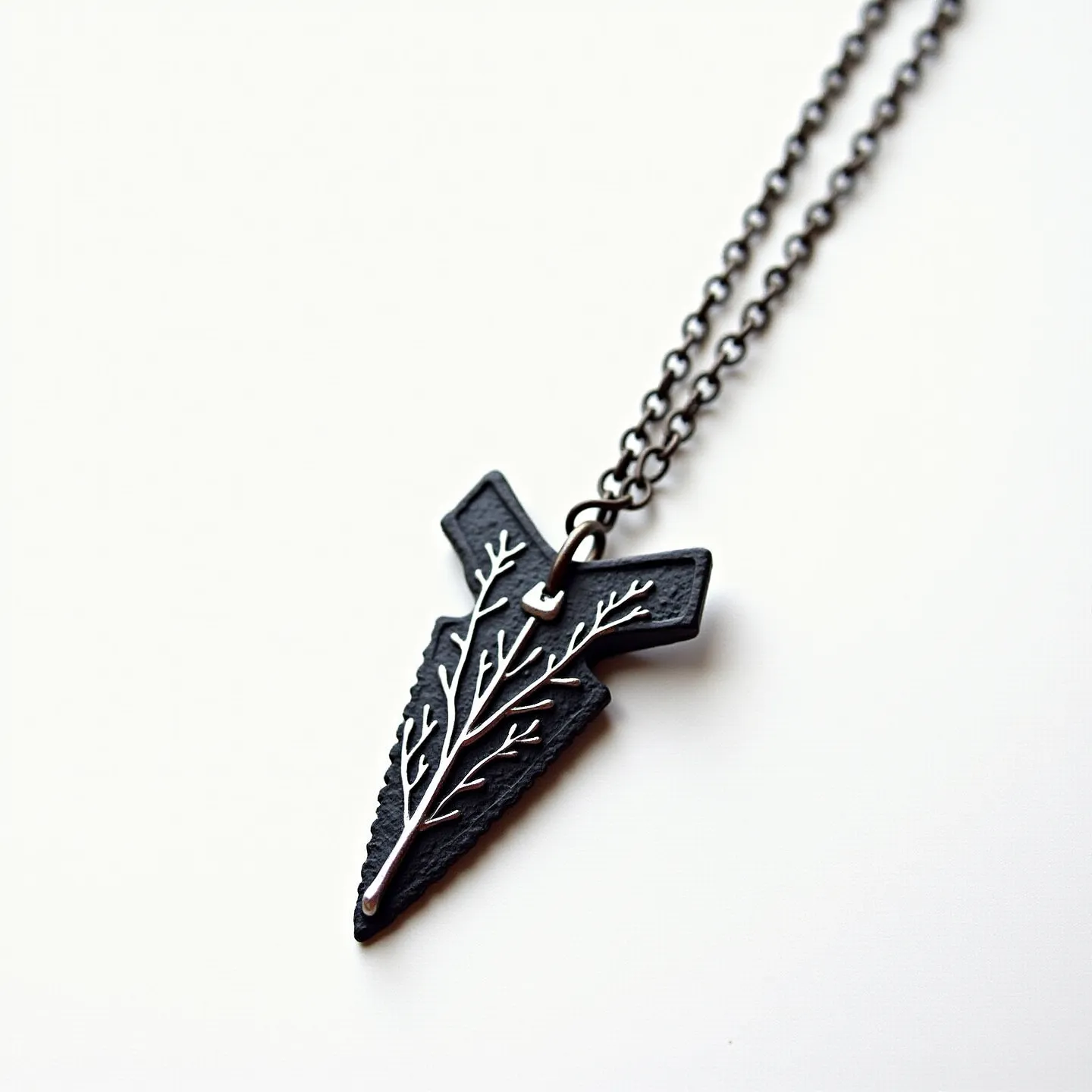 This arrowhead necklace features a dark, likely stone or metal pendant designed in the shape of an arrowhead. The surface is adorned with a delicate branching design, possibly crafted from a contrasting metal to enhance its visual appeal. The pendant is suspended from a chain composed of interlinked metal rings, providing a sturdy and complementary aesthetic. The necklace secures with a simple clasp or loop attachment, allowing for ease of wear.