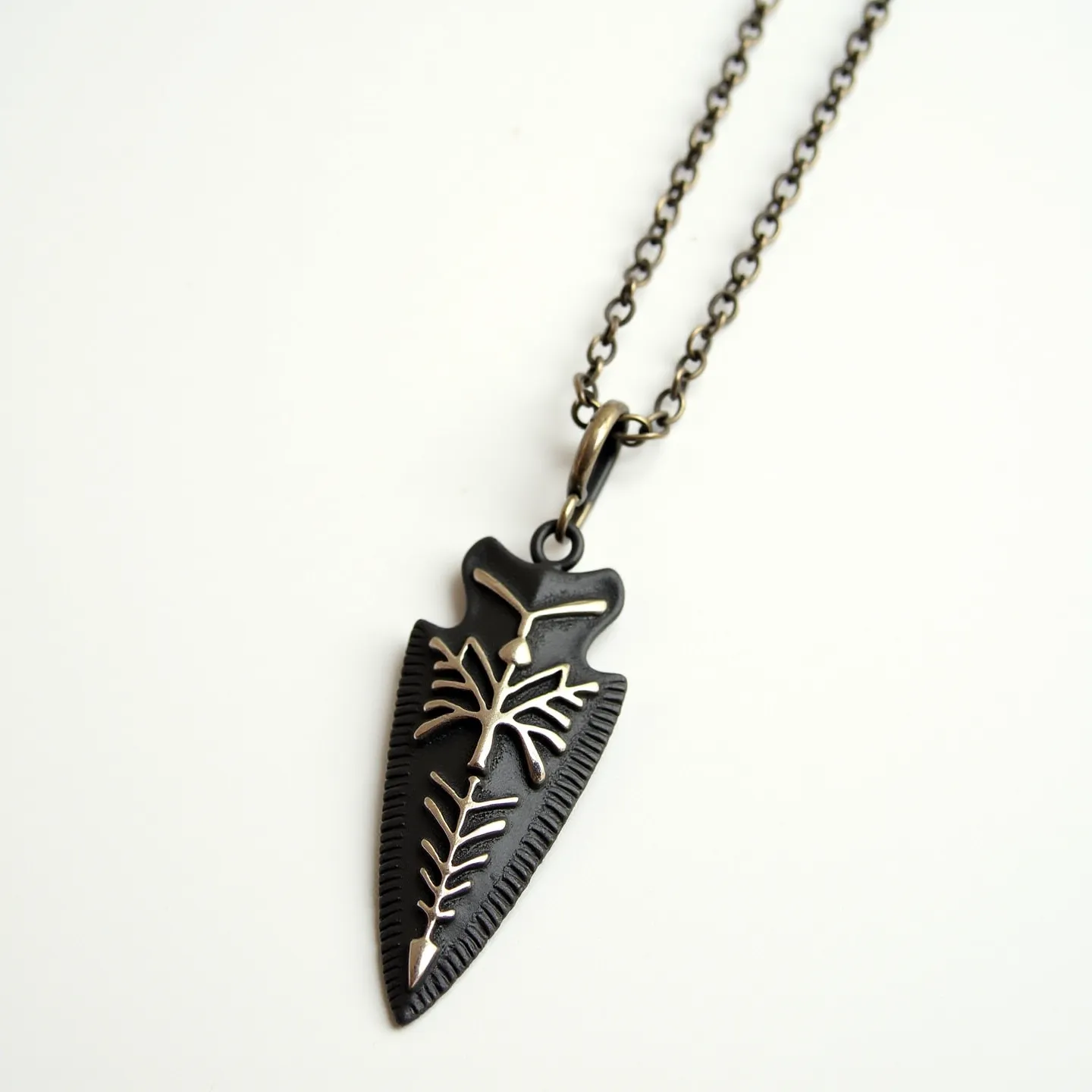 This arrowhead necklace features a striking design with a dark-toned metallic pendant shaped like an arrowhead, adorned with intricate silver-toned engravings that resemble tribal or natural motifs. The pendant is affixed to a robust, matching chain with a simple yet elegant ring clasp that ensures secure wear. The chain links are uniformly rounded, giving it a consistent and polished appearance. The combination of materials and detailed engravings gives this necklace a rugged aesthetic, enhanced by the contrasting finishes of the metals used for both the pendant and the chain.