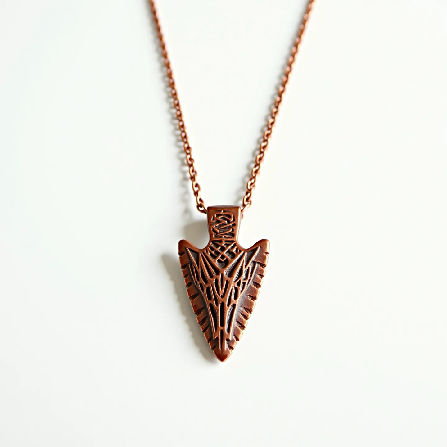 This arrowhead necklace features a pendant designed to resemble a stylized arrowhead, crafted from what appears to be a bronze material, giving it a rich, coppery hue. The pendant displays intricate, engraved patterns that add depth and texture to its surface. It is suspended from a chain that matches the pendant’s metallic tone, likely made from the same bronze material. The chain is composed of small interlocking links, which provide flexibility and movement. The connection between the pendant and the chain appears seamless, without any visible clasps or settings on the pendant itself, suggesting an integrated design that highlights the pendant as the focal point.