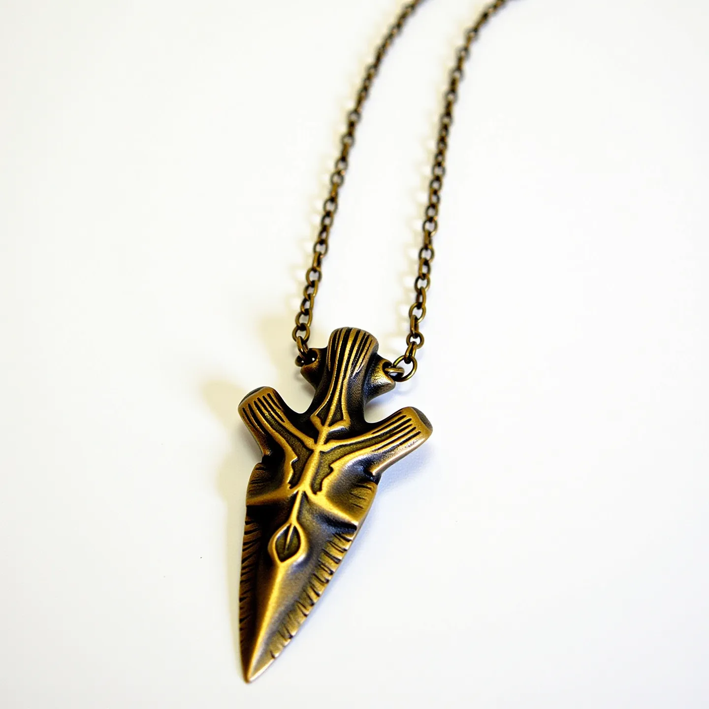 This arrowhead necklace features a metal pendant with a stylized design that suggests ornamental etchings and an aged finish. The pendant is connected to a bronze-toned chain, adding a rustic appeal to the piece. The arrowhead itself is crafted to resemble an ancient artifact, with symmetrical ridges and detailed engravings, but does not appear to include any gems. The chain attaches to the pendant through a small loop at the top, and likely secures with a standard clasp, though the clasp is not visible in this image.