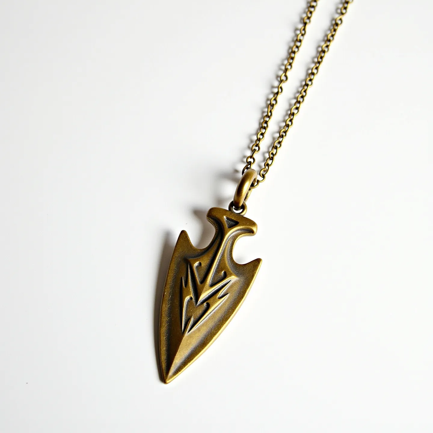 This arrowhead necklace features a pendant crafted from a brass-like material, showcasing a detailed, engraved arrowhead design with smooth, polished surfaces. The pendant is suspended from a simple link chain, also resembling brass, contributing to a cohesive aesthetic. The chain is secured with what appears to be a standard spring ring clasp, ensuring a reliable attachment. The overall design presents a bold, rustic charm, with the arrowhead's intricacy emphasizing a blend of artistry and symbolic significance.