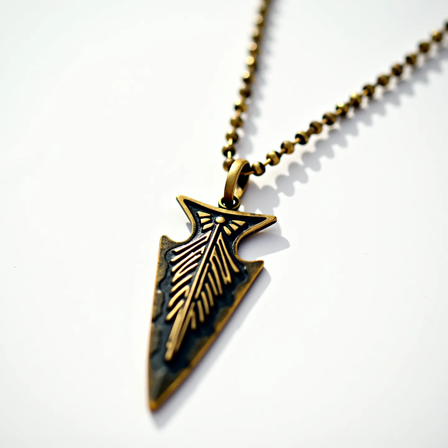 This arrowhead necklace features a metal pendant designed in the shape of an arrowhead with intricate engraved patterns on its surface, emphasizing its stylized form. The pendant appears to have an aged brass or antique gold finish, which gives it a vintage look. It is attached to a matching chain composed of small, interlinked round beads that complement the pendant. The necklace is equipped with a simple loop clasp that allows for secure fastening, harmonizing with the design of the chain and pendant.