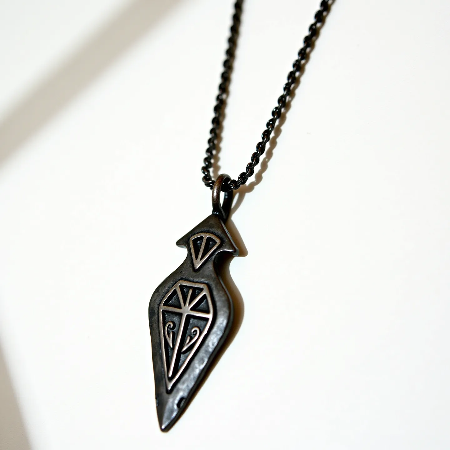 This arrowhead necklace features a pendant crafted from a dark metal, intricately designed with geometric and swirling patterns etched on its surface, giving it an artistic and ancient appearance. The pendant hangs from a twisted rope-style chain, which is also made of a similar dark material, adding to its cohesive and rugged aesthetic. The necklace is likely affixed with a simple loop attachment that seamlessly connects the pendant to the chain, allowing it to dangle freely and move naturally when worn. There are no evident gemstones or specialized cuts present in the design, maintaining a sleek and understated style. The overall impression of the necklace is one of bold simplicity, blending historical motifs with modern metalwork.