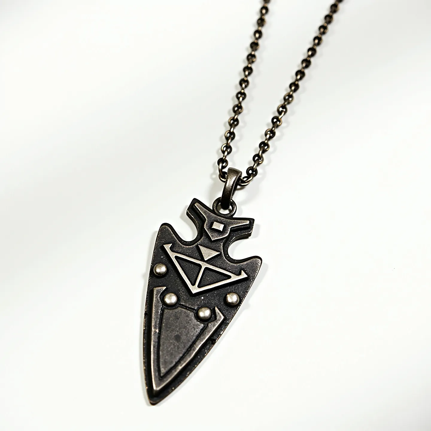 This arrowhead necklace features a dark metal pendant designed in the shape of an arrowhead. The pendant showcases geometric patterns and raised dots that add to its intricate design. It's attached to a dark metallic chain composed of small, round links, which gives it a robust appearance. The connection between the pendant and the chain is made through a sturdy loop at the top of the arrowhead, ensuring it hangs securely. The overall aesthetic is both bold and stylish, suitable for making a statement.