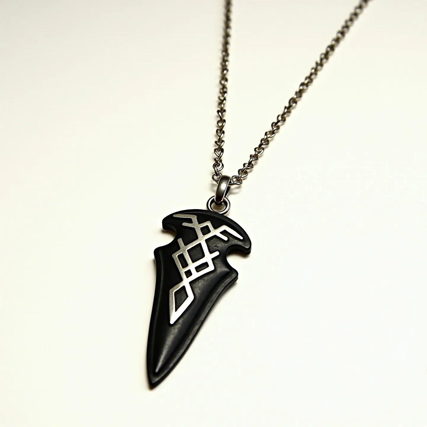 This arrowhead necklace features a striking pendant crafted from black metal or stone, shaped into an arrowhead with intricate geometric engravings. The design incorporates a silvery inlay that forms an elegant pattern across its surface, adding contrast and visual interest. The pendant is suspended from a robust chain with a dark metallic finish, enhancing the overall sleek and modern look. Attached to the pendant is a simple loop bail, through which the chain runs seamlessly. The necklace is secured by a standard lobster clasp, ensuring both ease of use and reliable closure.