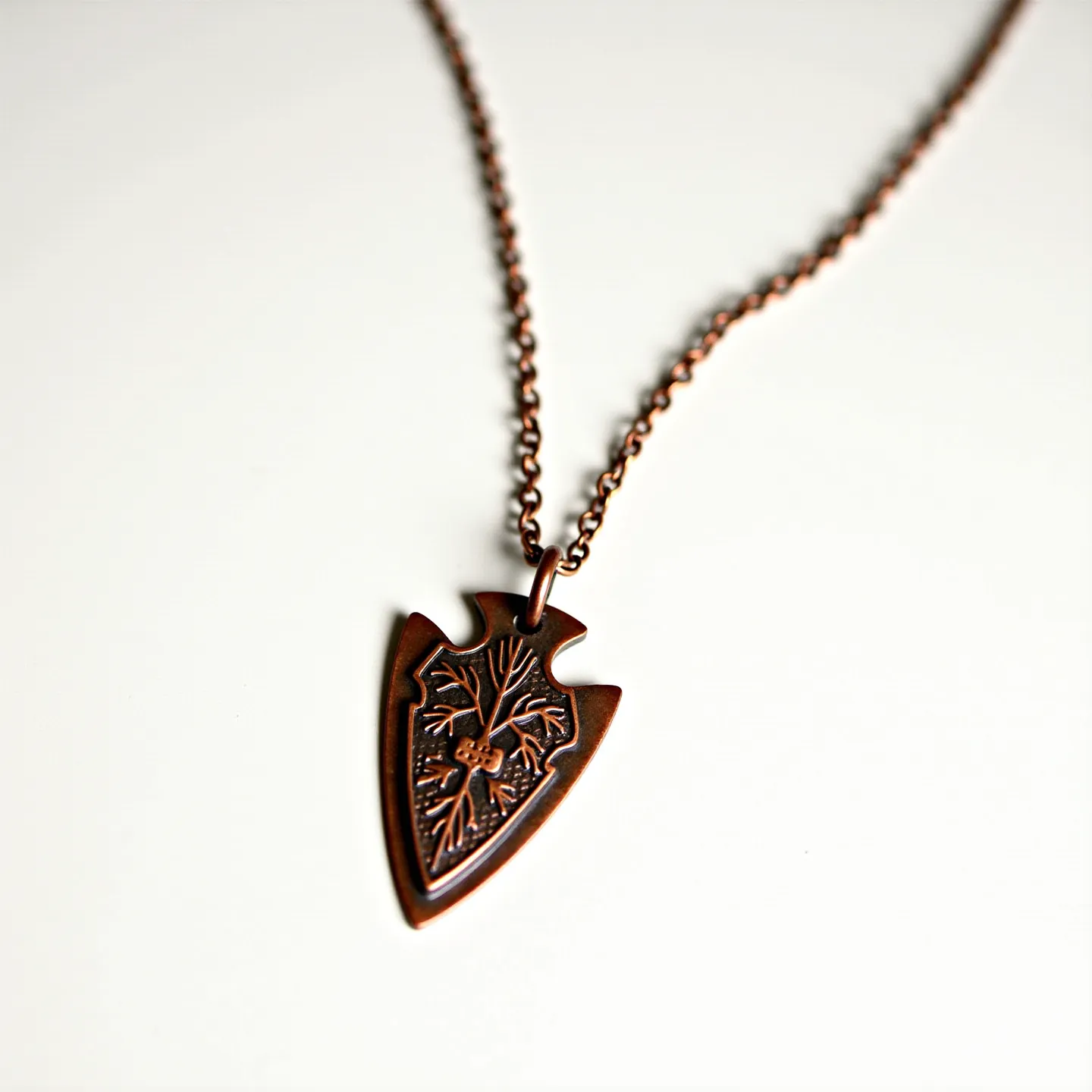 This arrowhead necklace features a pendant crafted from metal, with a copper finish that gives it a rustic and earthy appearance. The arrowhead shape is intricately detailed with engraved lines that suggest a natural, organic pattern, adding a unique visual interest to the piece. The pendant is attached to a matching copper-toned chain through a simple loop attachment, allowing for fluid movement. The chain resembles a cable link style, providing durability and complementing the overall aesthetic with its coordinated hue. A lobster clasp serves as the closure, ensuring secure wear while maintaining the necklace's cohesive design.