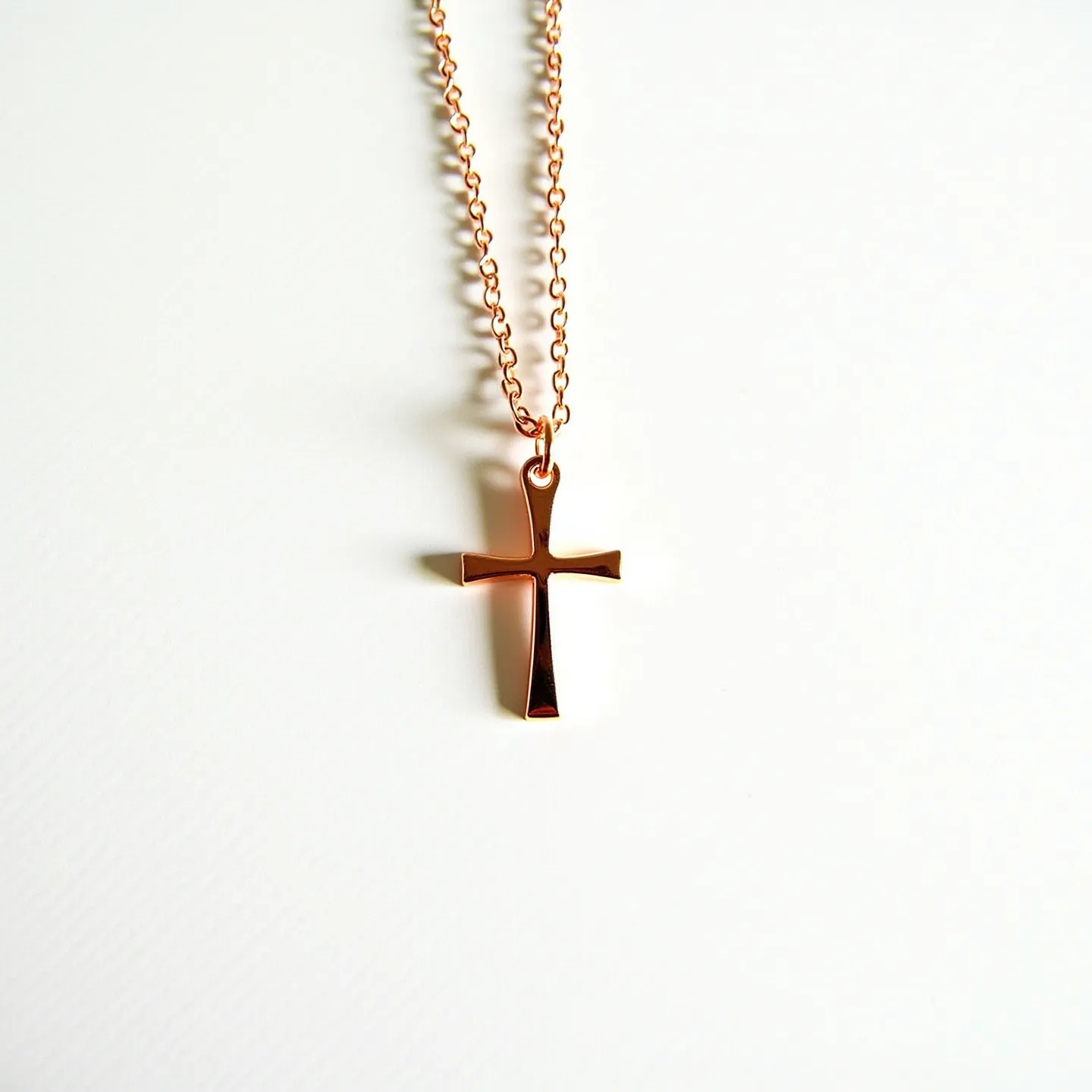 This baby cross necklace features a polished, rose gold-toned cross pendant with a smooth and reflective surface. There are no gemstones present on the pendant. The pendant is attached to a delicate chain via a small loop at the top of the cross, which seamlessly integrates into the design. The chain has a series of interconnected links that ensure flexibility and ease of wear. The simplicity and elegance of the design make it suitable for various occasions.