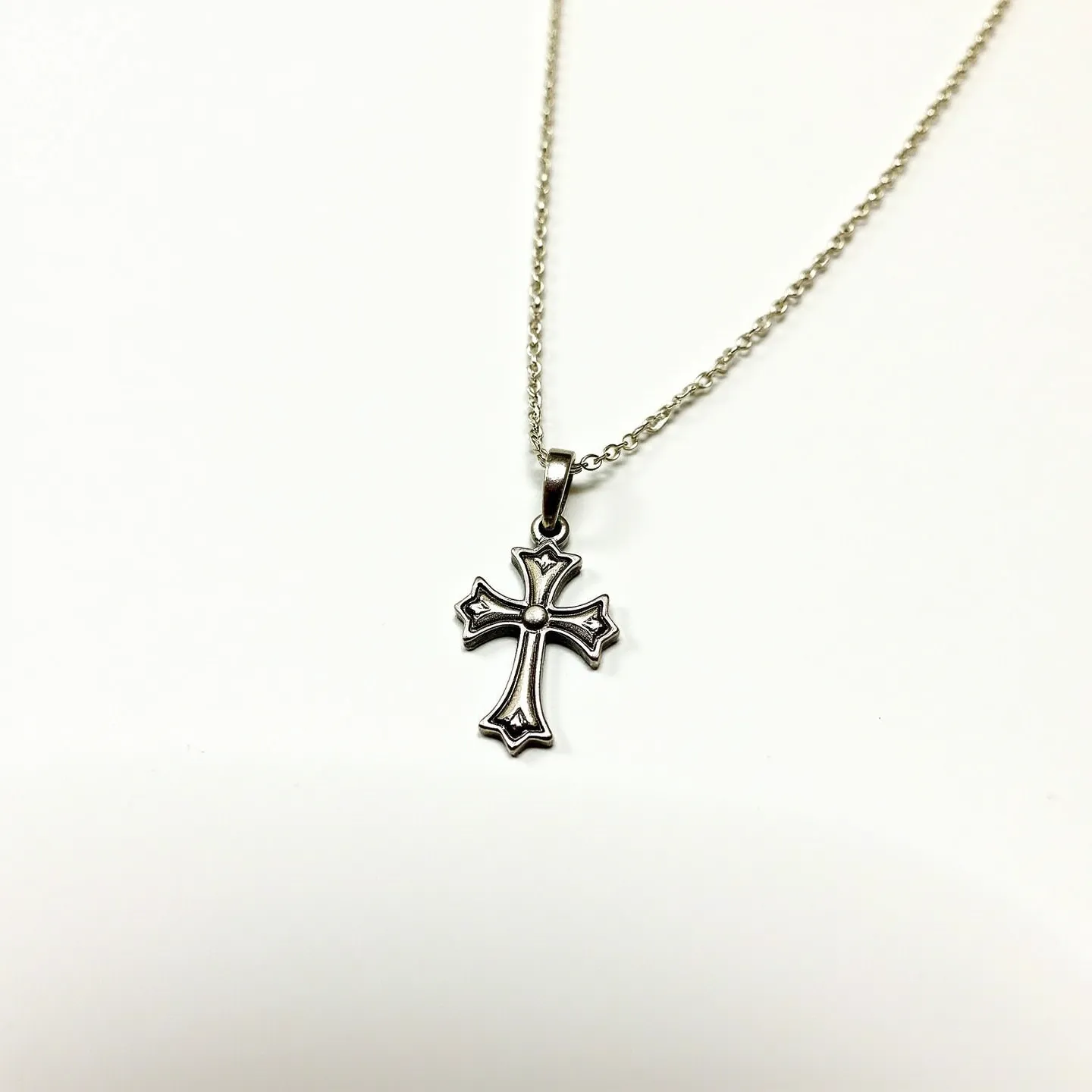 This baby cross necklace features a delicately designed cross pendant made of what appears to be a metal, likely silver or a silver-toned material, creating a bright and reflective finish. The pendant hangs from a fine link chain, which adds a subtle elegance to the piece. The cross itself is ornately decorated with intricate patterns that highlight its design. The necklace is secured with a small, simple ring attachment at the top of the cross, allowing it to move freely along the chain and ensuring ease of wearing. The overall craftsmanship suggests a piece that is both stylish and suitable for a baby or a special occasion.