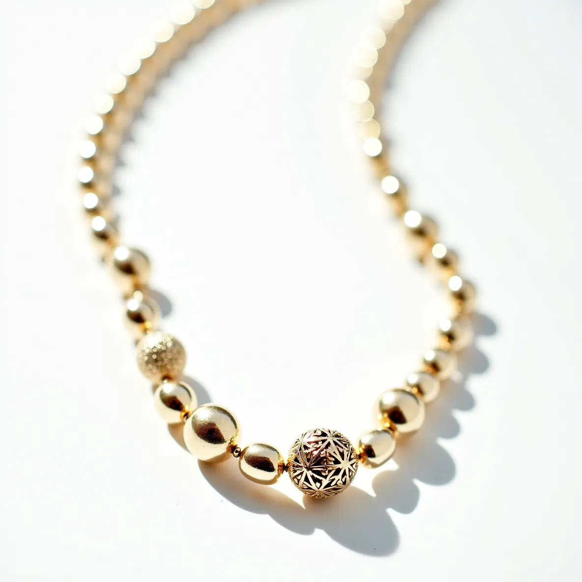 This beaded necklace features a series of smooth, gold-toned spherical beads, interspersed with a few uniquely designed focal beads. One bead showcases an intricate filigree pattern, adding an element of elegance and complexity to the design. Another bead is textured, providing a contrast to the smooth finish of the surrounding beads. A variety of bead sizes enhances the visual interest of the piece. The necklace is strung together on a uniform, durable string, likely possessing a clasp or other attachment for secure fastening, though it is not visible in this view. The overall design suggests a sophisticated and versatile accessory suitable for both casual and formal occasions.