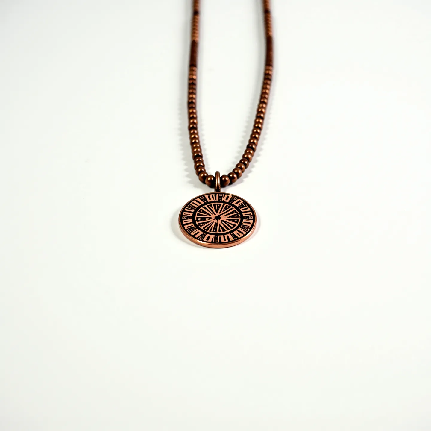 This beaded necklace features a series of small, evenly spaced beads that appear to be made of a shiny, metallic material, likely copper or a copper-colored alloy, given their reflective, warm hue. Suspended from the center of the necklace is a circular pendant, also in a coppery tone, adorned with intricate, engraved designs that suggest a sun or wheel motif. The pendant is attached to the necklace with a loop that matches the beaded material, providing a cohesive and harmonious design. The necklace seems to have a minimalist approach, focusing on the natural sheen and color of the metallic beads and pendant.