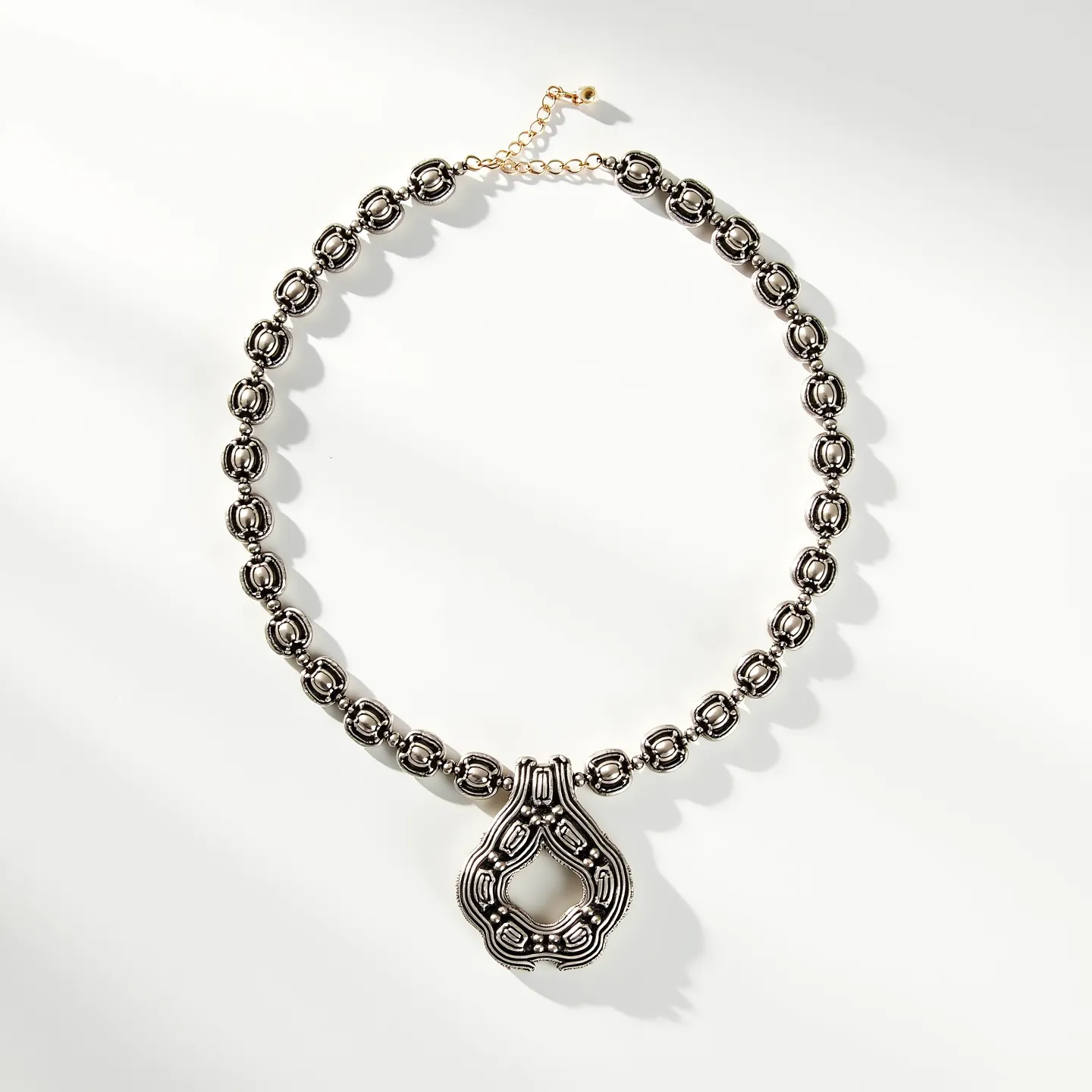 This beaded necklace features metallic beads with an intricate design, appearing to be crafted from silver or a silver-toned metal. The beads are uniform, creating a rhythmic pattern throughout the necklace. At the center hangs a prominent pendant with a hollow, teardrop-like shape, echoing the beaded design's motifs. The clasp is a delicate chain extension with a small bead at the end, likely adjustable to accommodate different lengths when worn.