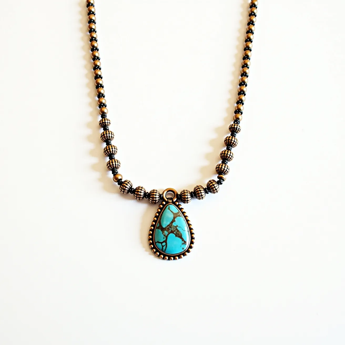 This beaded necklace features a series of metallic beads, alternating between smooth and ornately textured designs. It centers around a striking teardrop-shaped turquoise stone, characterized by its vibrant blue color with dark veining patterns. The turquoise is held in place by a detailed bezel setting with a decorative border that complements the beads. The necklace is completed with a loop attached to the pendant, seamlessly blending with the overall design and ensuring a secure attachment.