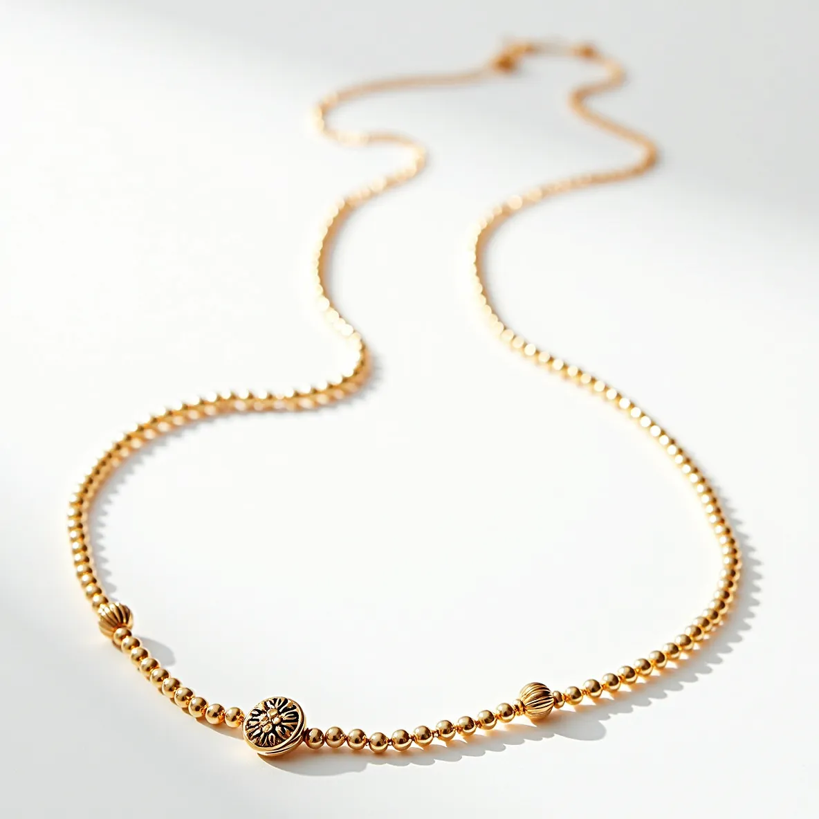 This beaded necklace features small, round beads with a gold tone, interspersed with occasional larger, intricately detailed beads that add visual interest and texture. Central to the design is a prominent bead with a sunburst pattern that serves as a focal point. The sunburst bead appears to be engraved or embossed, contributing to the necklace's decorative appeal. The necklace is strung on a thread or chain that ensures flexibility and ease of wear. For closure, it appears to have a standard hook clasp, providing a secure attachment. The overall design displays a harmonious balance of simplicity and ornate detail, making it suitable for both casual and formal occasions.