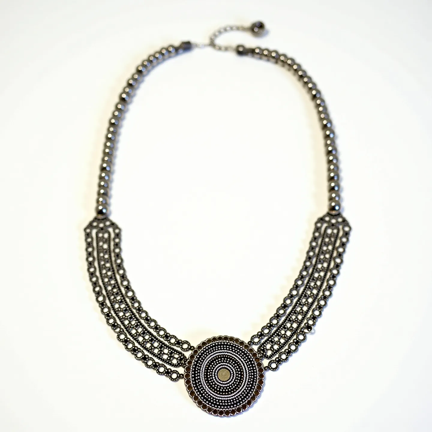 This beaded necklace features intricately arranged small, dark metallic beads that form a symmetrical design. The central focal point is a large circular pendant, crafted with meticulously layered concentric patterns of beads. The beads appear to be metallic, likely made of hematite or a similar material, giving the piece a cohesive and monochromatic look. The necklace culminates in a straightforward clasp and extender chain, allowing for adjustable length and ease of wear. The overall aesthetic is both elegant and striking, making it suitable for various occasions.