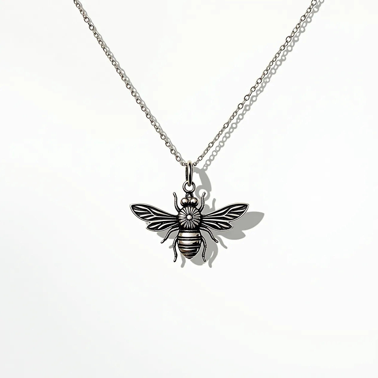 This bee necklace features a detailed bee pendant crafted from a metallic material, likely silver due to its sheen and color. The pendant showcases intricate engraving that highlights the bee's wings and body segments, creating a textured appearance. It is suspended from a delicate chain with small, evenly spaced links. There are no visible gems or stones set into the pendant, and the overall design emphasizes the artistry of the metalwork. The necklace is equipped with a simple loop at the top of the bee connecting to the chain, likely ending in a standard clasp for secure fastening.