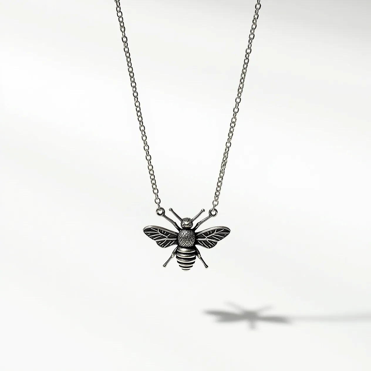 This bee necklace features a detailed bee pendant crafted from a silver-toned metal. The pendant is intricately designed to highlight the bee’s wings, body, and antennae, giving it a textured appearance. The necklace is suspended on a delicate chain that complements the pendant's metallic finish. The chain is likely secured with a standard clasp, ensuring it can be easily opened and closed while worn. There are no visible gemstones or embellishments, allowing the design to focus on the elegant form of the bee itself.