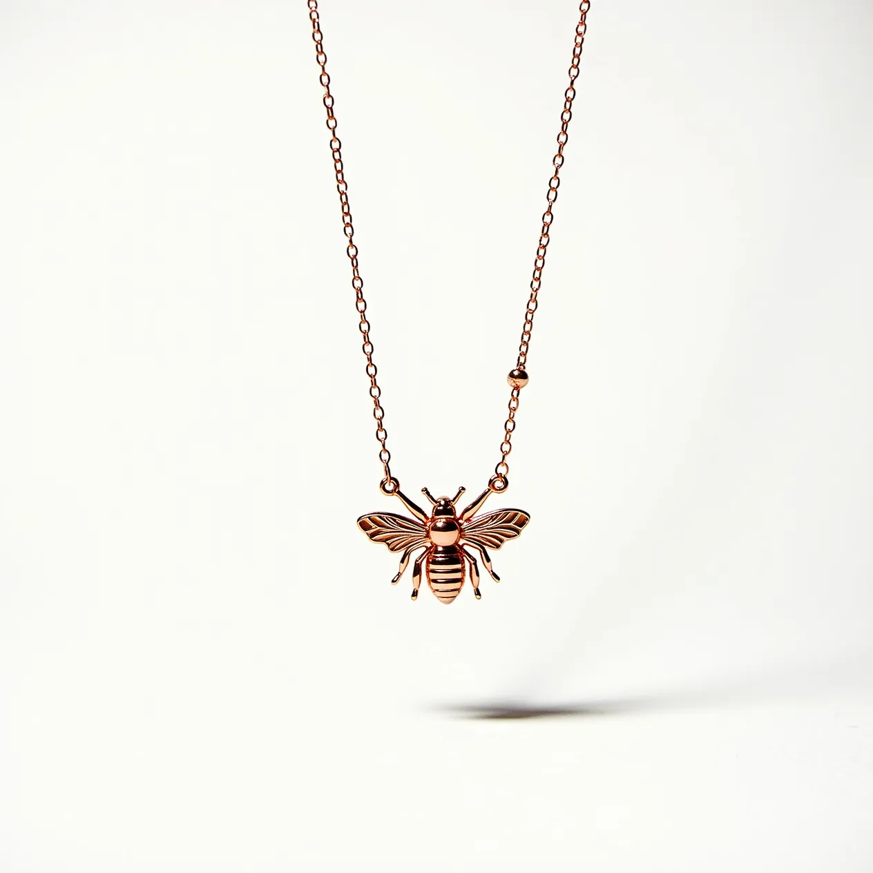 This bee necklace features a charming bee-shaped pendant made of a warm, rose gold-toned metal, intricately detailed to capture the delicate features of the insect. The pendant hangs from a fine chain that matches its metallic hue, adding to the cohesive look of the piece. The necklace includes a small, decorative bead integrated into the chain, adding a touch of understated elegance. Additionally, the necklace has a standard clasp for secure attachment and easy wearability. The entire design combines simplicity with a natural motif, making it a stylish accessory.