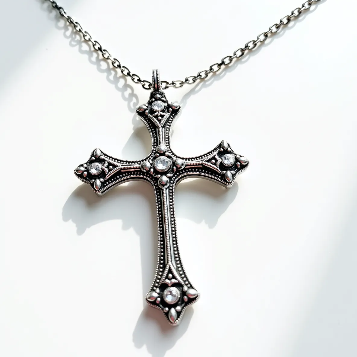 This big cross necklace features a detailed design crafted from a metal that gives it a silver-like appearance. The cross is adorned with clear, round-cut gems set in the center and at each of the four ends, adding a touch of sparkle and elegance. The metal is intricately detailed with beading along its edges, enhancing its ornate and classic look. The pendant is suspended on a medium-length chain with interlocking links, which is likely metal as well, and includes a small loop at the top of the cross for attachment. This loop connects seamlessly to the chain, ensuring the pendant is securely displayed.