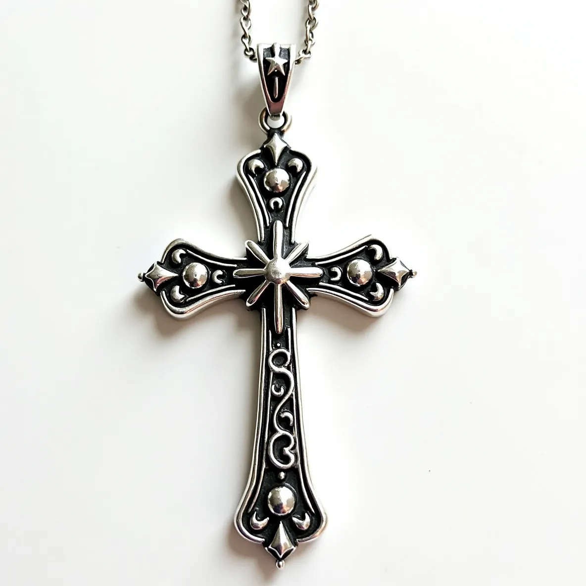 This big cross necklace features an ornate design crafted from a metallic material with intricate detailing along its surface. Each arm of the cross is accented with embellishments resembling polished round beads or studs, enhancing its decorative appeal. At the center, a starburst motif adds further dimension, creating a focal point. The pendant is attached to a chain via a bail featuring a small star detail. The chain itself appears to have an interlinked design, providing both strength and style. Overall, the piece combines classic religious symbolism with elaborate artistic elements.