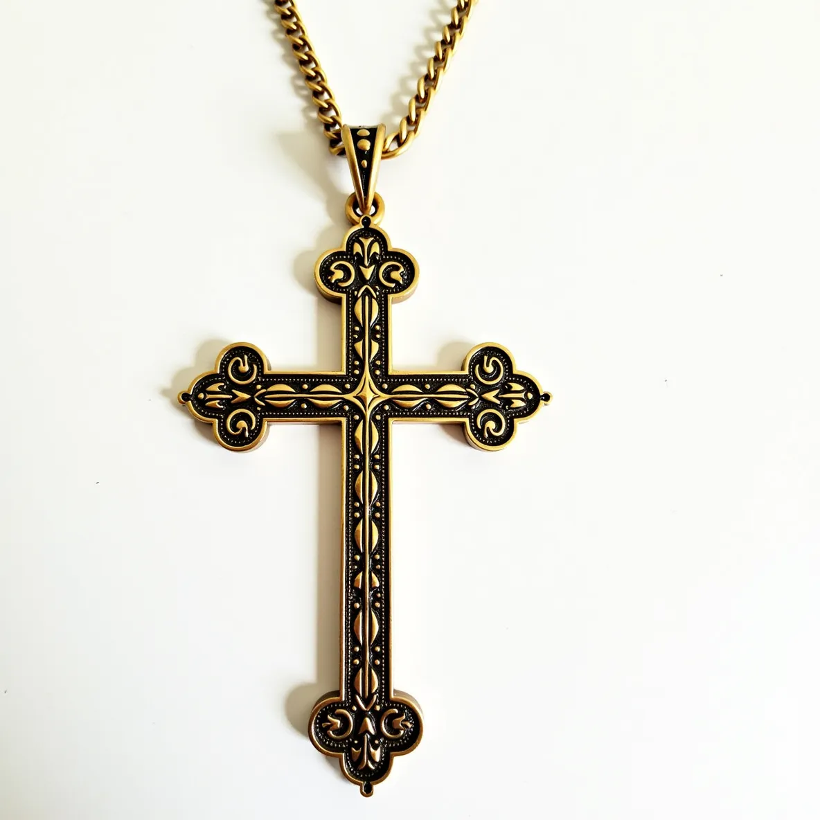 This big cross necklace features an intricate, ornate design crafted from a metal with a rich, gold finish, possibly brass or gold-plated. The cross exhibits detailed patterns, giving it a vintage or gothic aesthetic. The pendant appears to be large in size, with decorative elements enhancing its visual appeal. The cross is suspended from a matching gold-tone chain with a traditional link pattern. The chain is attached to the cross via a decorative bail, which seamlessly connects the pendant to the chain, allowing it to be worn comfortably. The clasp is not visible, but it likely involves a standard fastener suitable for this type of necklace.