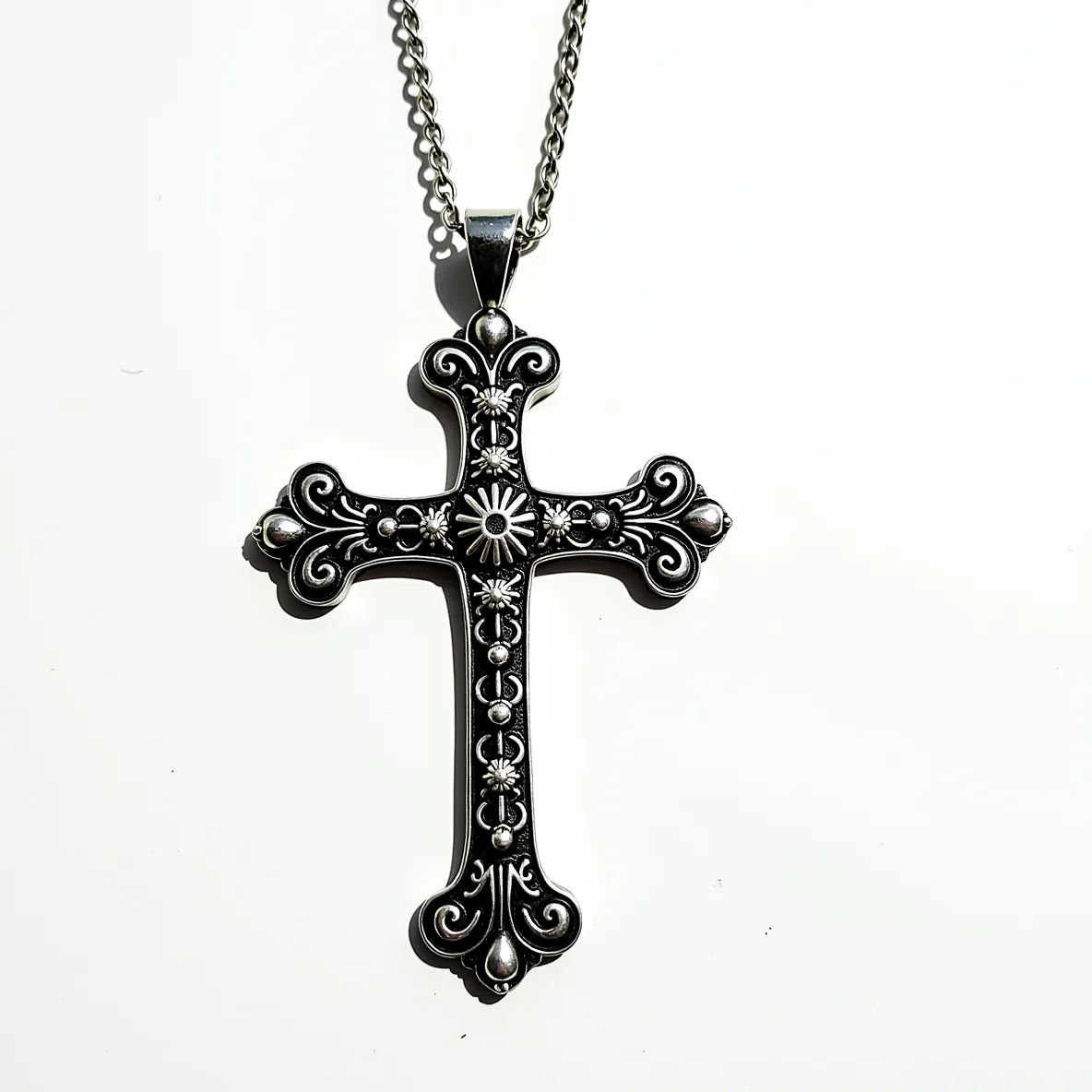 This big cross necklace features an intricately designed metal pendant, likely made of silver or a similar metal, with ornate patterns and detailing. The surface showcases raised dots and floral motifs that enhance its decorative appeal. At each end of the cross, rounded embellishments add dimension and texture. The pendant is suspended from a medium-thick chain, suggesting durability and sturdiness, and is attached through a substantial bail that allows for a fluid movement with the chain. The overall design hints at a vintage or gothic style, making it both a statement piece and an accessory with a classic aesthetic.