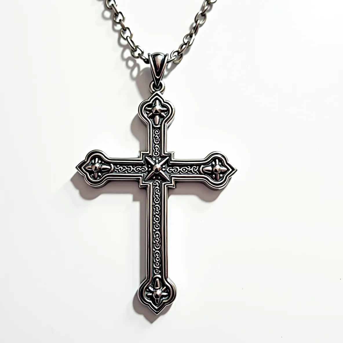 This big cross necklace features an intricate silver metal design with ornate embellishments. The cross showcases detailed engravings along its surface, with raised motifs that may resemble small floral patterns at each endpoint. At the center, a star-like design adds dimension to the piece. The cross is attached to a robust silver chain, and the attachment is a solid bail that connects the pendant securely to the chain. The overall design suggests a gothic or vintage-inspired style, highlighting the craftsmanship of the intricate detailing.