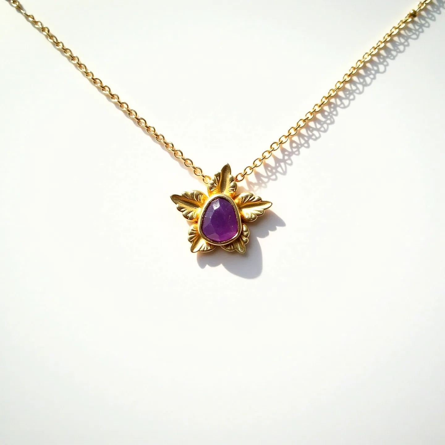 This birthstone necklace features a gold-toned chain supporting a pendant with a polished purple gemstone, likely amethyst, set in a bezel setting. The gem is encircled by a decorative gold leaf design that enhances its elegance, offering a vintage charm. The chain is composed of interlocking links, which provide durability and a classic aesthetic. An attached clasp facilitates easy wear while ensuring security. The necklace's design showcases a balance of simplicity and sophistication, making it a versatile accessory.