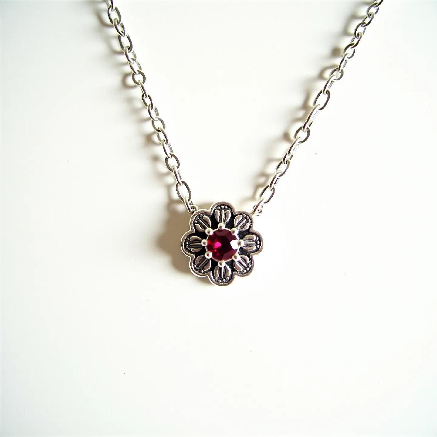 This birthstone necklace features a delicate chain made of a polished metal, likely silver, which supports a striking pendant. The pendant itself has an intricate floral design, crafted with detailed metalwork that adds a vintage touch. At its center, the necklace showcases a brilliant red gemstone, possibly a ruby or garnet, cut into a round shape and securely held in place by four metal prongs in a classic prong setting. The necklace includes a standard clasp, ensuring it can be easily fastened and worn securely.