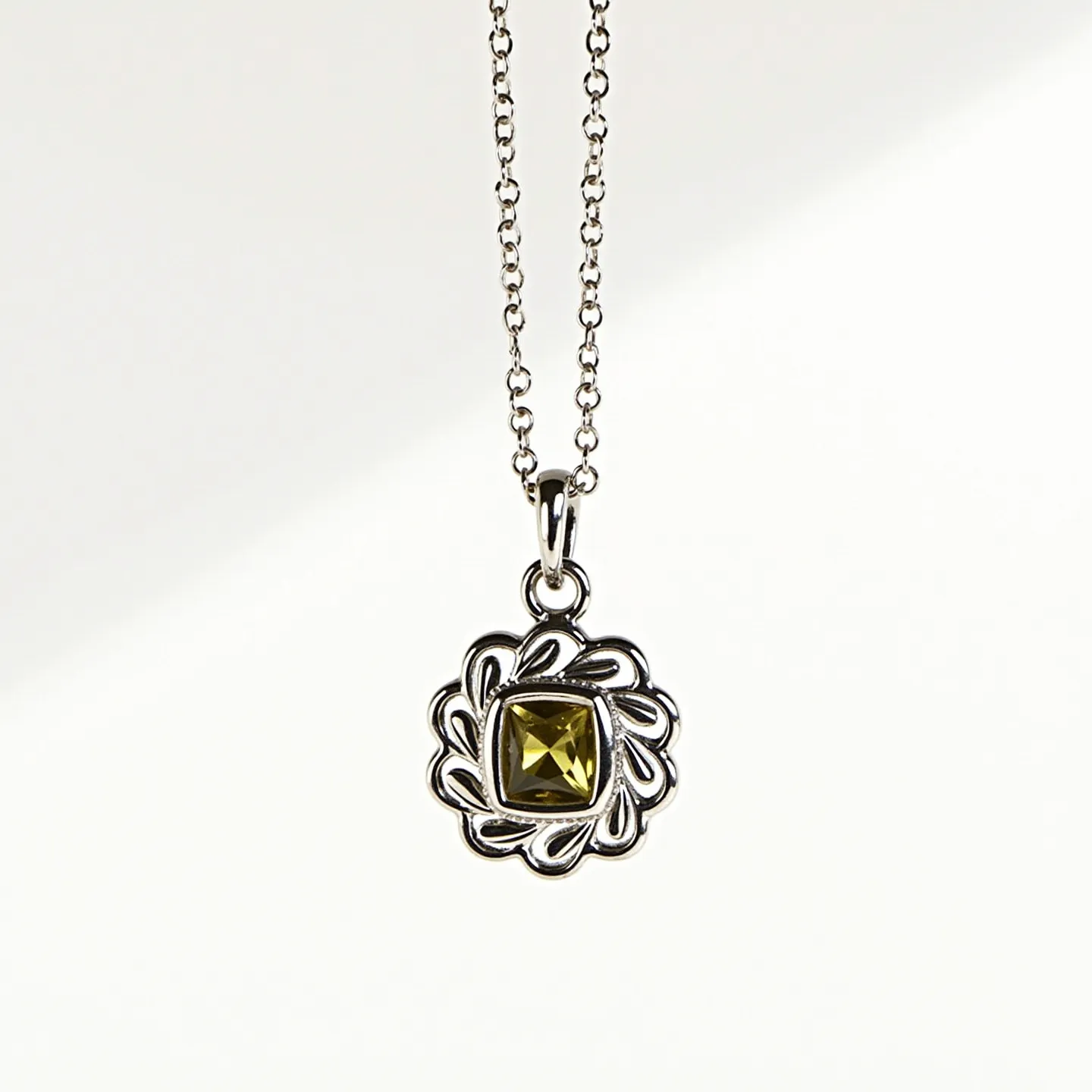 This birthstone necklace features a striking square-cut gemstone, possibly a peridot, set in a decorative, ornate metal setting with a floral or abstract swirl design. The metal is likely silver or a silvery alloy, providing a polished and elegant contrast to the stone's vibrant green hue. The pendant hangs from a delicate chain, which is composed of small, linked circles, enhancing the overall graceful appearance of the necklace. The chain is attached to the pendant with a simple bail, allowing for fluid movement and a classic look.