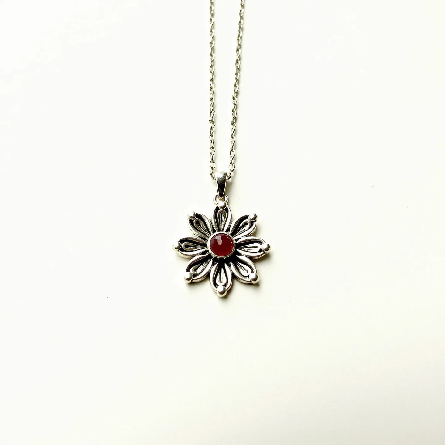 This birthstone necklace features a beautifully crafted silver pendant shaped like a flower with intricate petal designs. At the center is a round, polished red gemstone, possibly a garnet, which is commonly associated with the month of January. The gemstone is securely set in a bezel setting, enhancing its elegant appearance. The necklace is attached to a fine silver chain with a delicate and secure bail, providing both style and durability. The overall design is simple yet sophisticated, making it a timeless piece of jewelry.