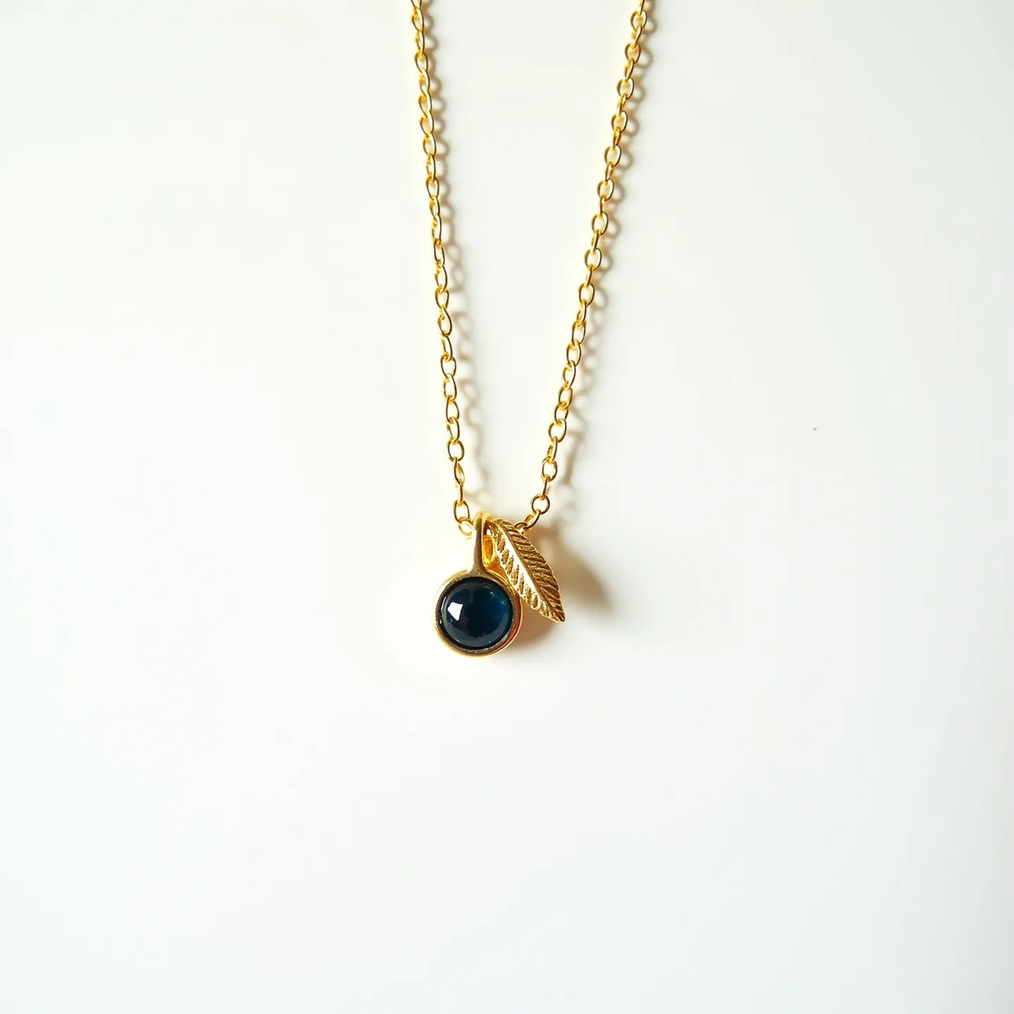 This birthstone necklace features a delicate gold chain, showcasing a deep blue gemstone set in a simple bezel setting. The stone, which could be a sapphire or similar gem, is round with a smooth cabochon cut, adding a classic elegance to the piece. Accompanying the gemstone is a small gold leaf charm, intricately detailed to complement the overall design. The necklace is secured with a standard clasp, allowing for easy wear and removal while maintaining a secure fit.