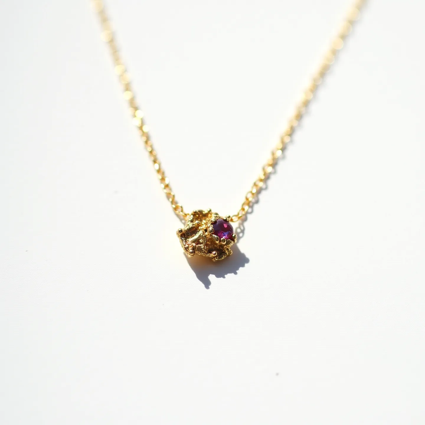 This birthstone necklace features a delicate gold chain with a textured gold nugget pendant. Set within the nugget is a small, round-cut gemstone that appears to be a deep red hue, likely representing a ruby. The stone is held securely in place, allowing its vibrant color to catch the light. The necklace is completed with a subtle chain design that enhances its elegant appearance.