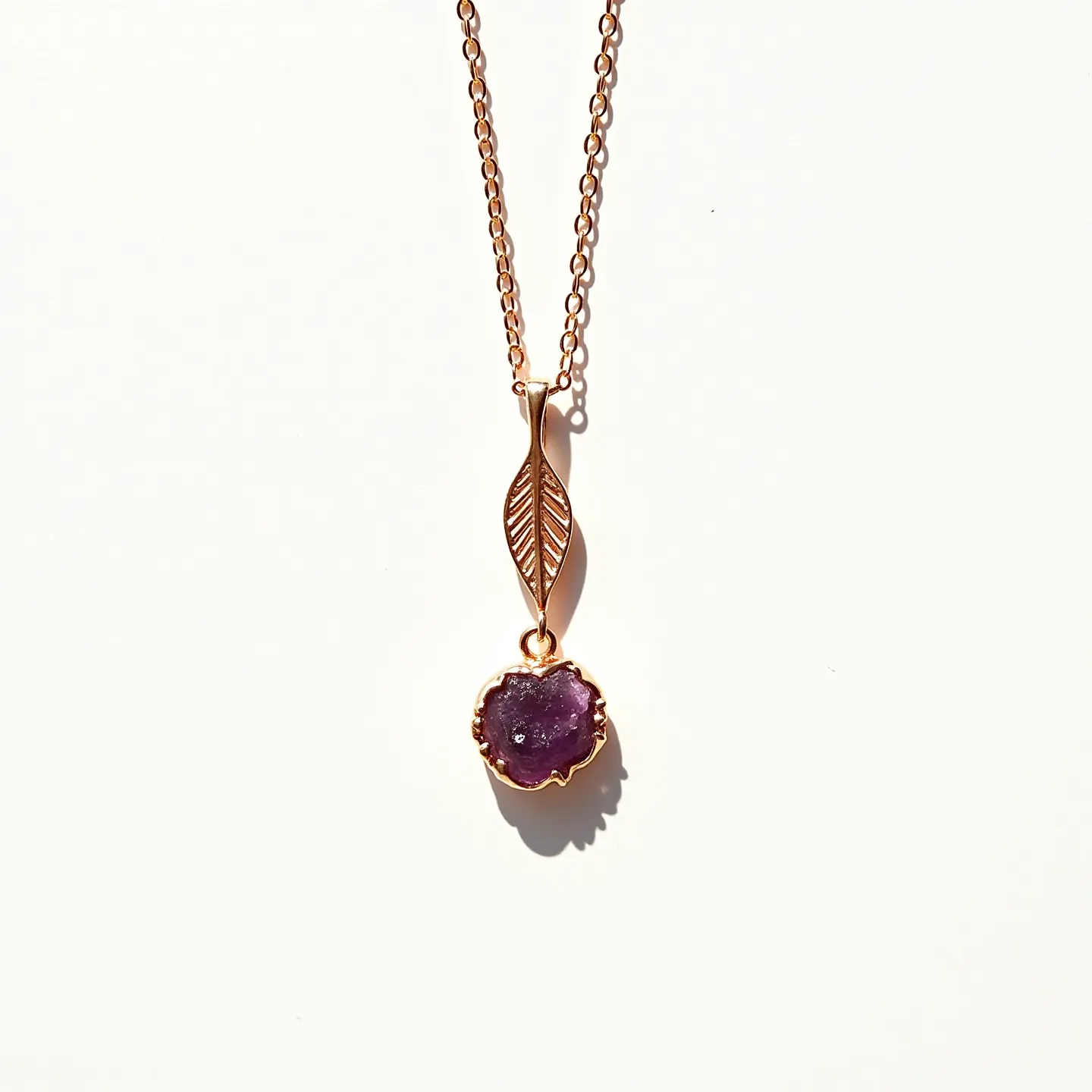 This birthstone necklace features a delicate chain crafted from a warm-toned metal, likely gold or gold-plated. Suspended from the chain is a uniquely shaped pendant that includes a leaf motif, giving the piece a natural and organic aesthetic. Below the leaf design hangs a stunning raw amethyst gemstone, identifiable by its deep purple hue and irregular, organic cut. The stone is securely set in a scalloped metal bezel that complements the overall design with its earthy elegance. The necklace likely has a standard clasp at the back for secure wearing.