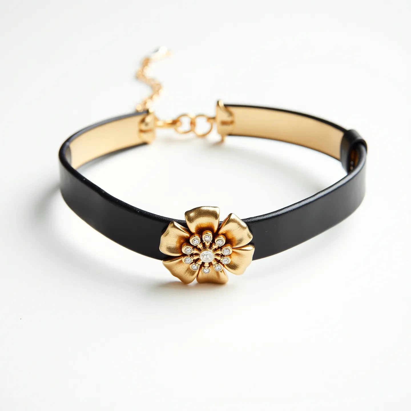 This black choker features a sleek band, likely made from a smooth leather or satin-like material, giving it a refined appearance. At its center is a striking floral embellishment crafted from metal with a gold-tone finish, designed to resemble a flower with layered petals. The center of the flower holds multiple small, clear stones, possibly cubic zirconia or crystals, stylishly set in a cluster that mimics a classic pave setting, adding a touch of sparkle. The choker is equipped with an adjustable chain clasp, which is also gold-toned, providing both functionality and an element of elegance.