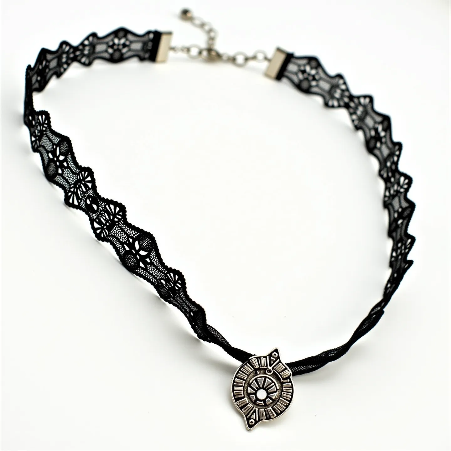 This black choker features a delicate lace material with intricate floral patterns running along its length. At the center, it is adorned with a small, round pendant, which appears to be metallic with detailed engravings. The choker is adjustable and secured by a lobster clasp accompanied by a chain extender, allowing for varying fits. The overall design is both elegant and versatile, suitable for a range of styles.