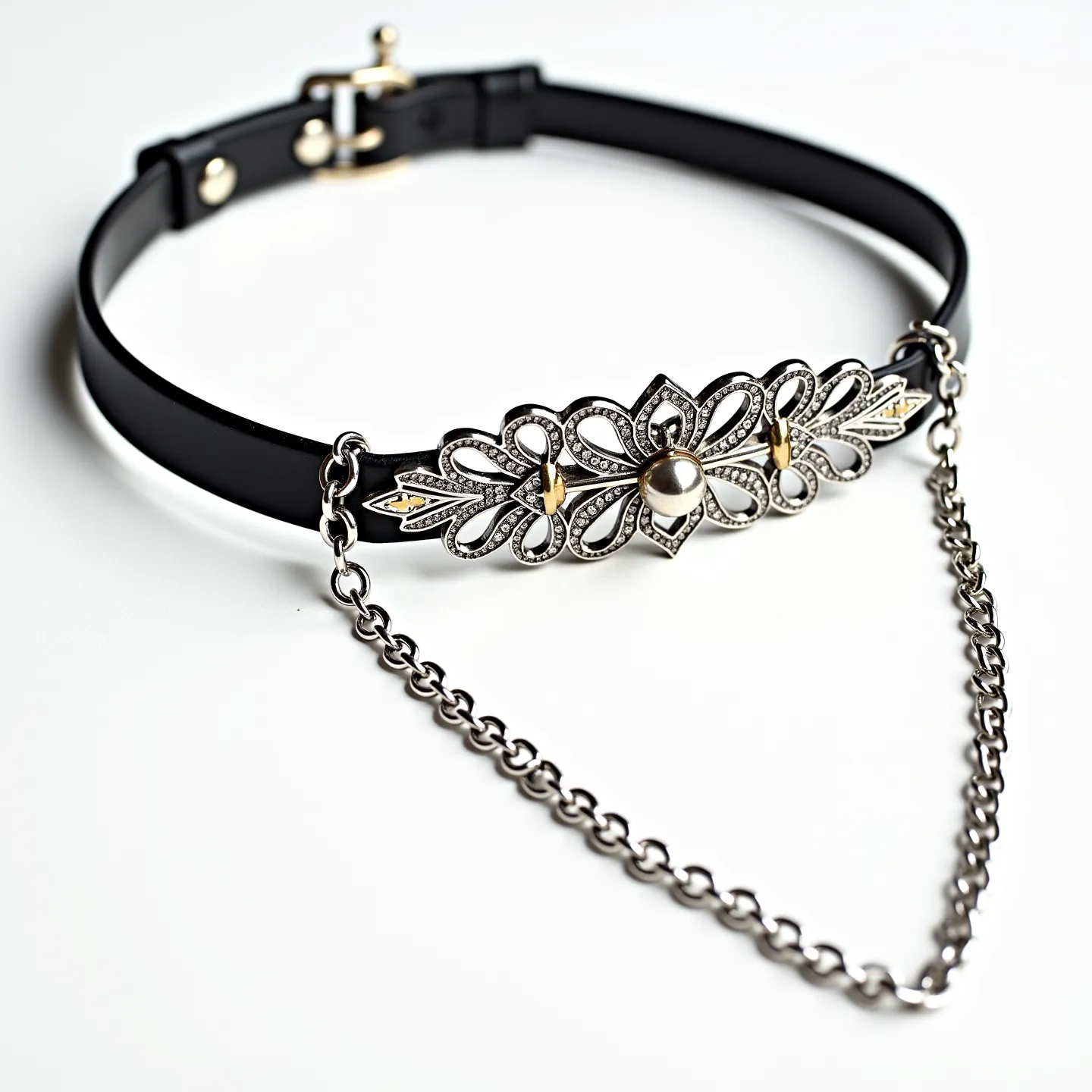 This black choker features a sleek band made of likely synthetic or leather material, providing a smooth and elegant backdrop. The centerpiece is an ornate metallic design, intricately shaped with cut-out patterns and accented with small embellishments for added texture. The embellishments include a central rounded gem, possibly a pearl, set prominently amidst the metallic flourishes. Additionally, two gold-toned accents flank the centerpiece, adding contrast and dimension. The choker is secured by a metallic clasp resembling a tiny buckle, ensuring a snug fit, complemented by a decorative chain draping partially across the front for added flair.