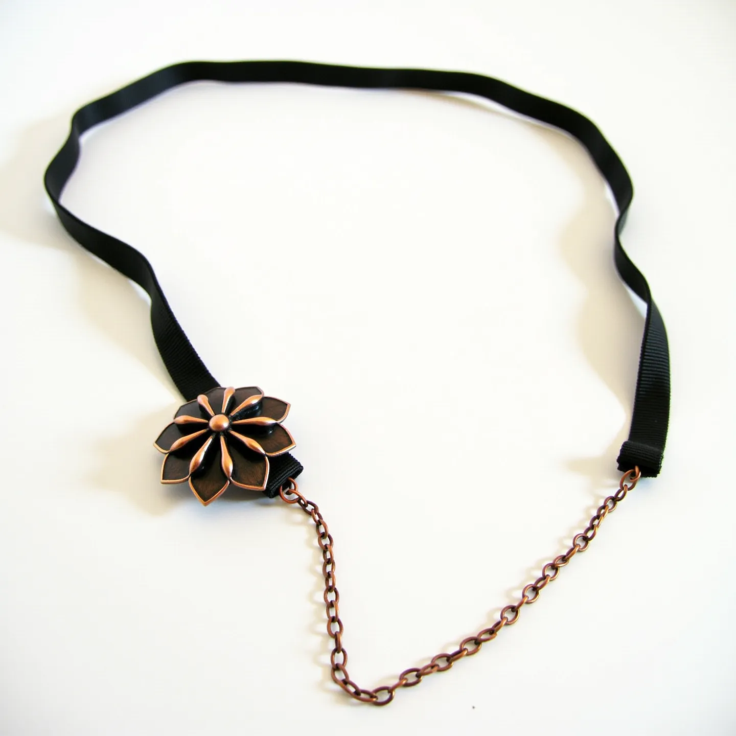 This black choker features a delicate, flat black ribbon as its main component, creating a sleek and simple base. The centerpiece is an ornate bronze flower design, with multiple petal layers extending from a central raised point, giving it an elegant and vintage-inspired appearance. The petals have a polished finish, augmenting the intricate design with subtle shine. The choker is completed with a bronze-colored chain that adds an additional decorative touch and likely serves as a fastening mechanism, providing an adjustable fit around the neck. The combination of the black ribbon and bronze elements creates a striking contrast, enhancing the choker's overall aesthetic.