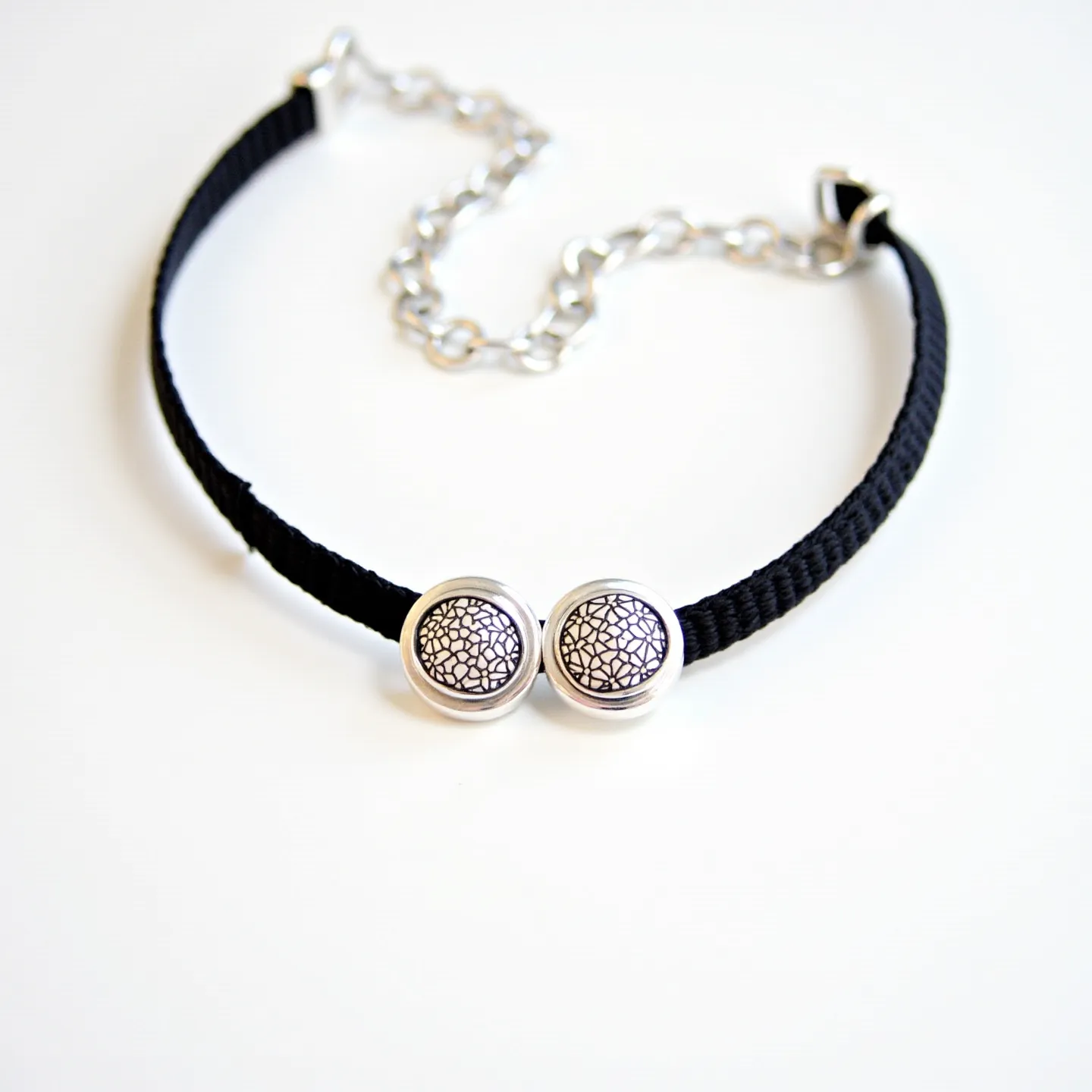 This black choker features a braided or woven band, likely made from a soft fabric or similar material. At the front of the choker, there are two round decorative elements with intricate mosaic patterns encased in silver-tone settings, providing a touch of elegance. The choker is adjustable with a silver-tone chain and secured by a lobster clasp, allowing for comfortable and customizable wear. The combination of the black band and silver elements creates a classic and versatile accessory suitable for various styles.