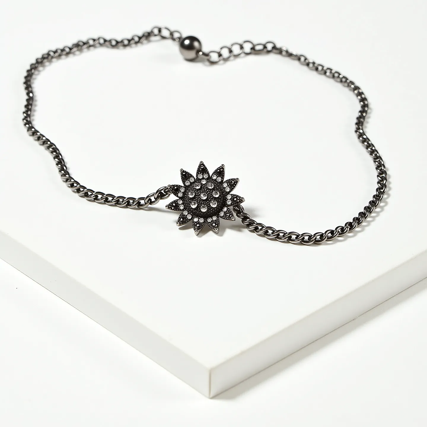 This black choker features a chain made of a dark, possibly oxidized metal, creating a sleek and elegant look. At the centerpiece is a striking floral motif adorned with numerous small, round black stones that are likely set in a pavé style, adding texture and sparkle. The stones create a distinctive, eye-catching design reminiscent of a sunflower. The choker is equipped with an adjustable clasp, allowing for flexibility in fit, which is typical with ball-and-chain style attachments for a secure closure.