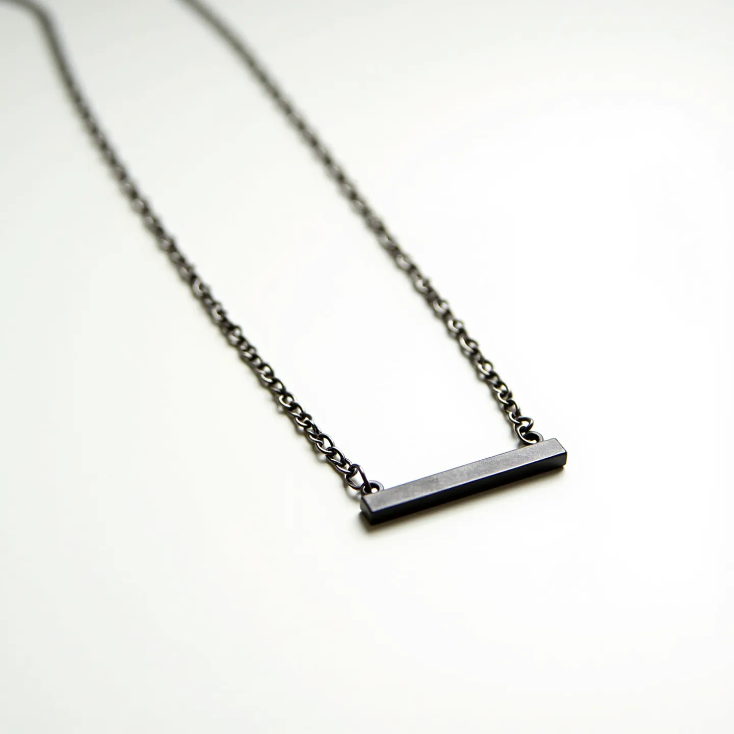 This boy's chain necklace features a simple yet striking design with a sleek, dark metallic finish. The necklace consists of a series of small, uniformly interlinked chain links that give it a slightly industrial appearance. Suspended at the center is a rectangular bar pendant, also in the same dark metal, adding a contemporary touch to the piece. The clean lines of the bar pendant provide a minimalist aesthetic, with no additional embellishments or gemstones. The attachment and clasp are not visible in this particular view, but the overall design suggests a focus on modern simplicity and understated style.