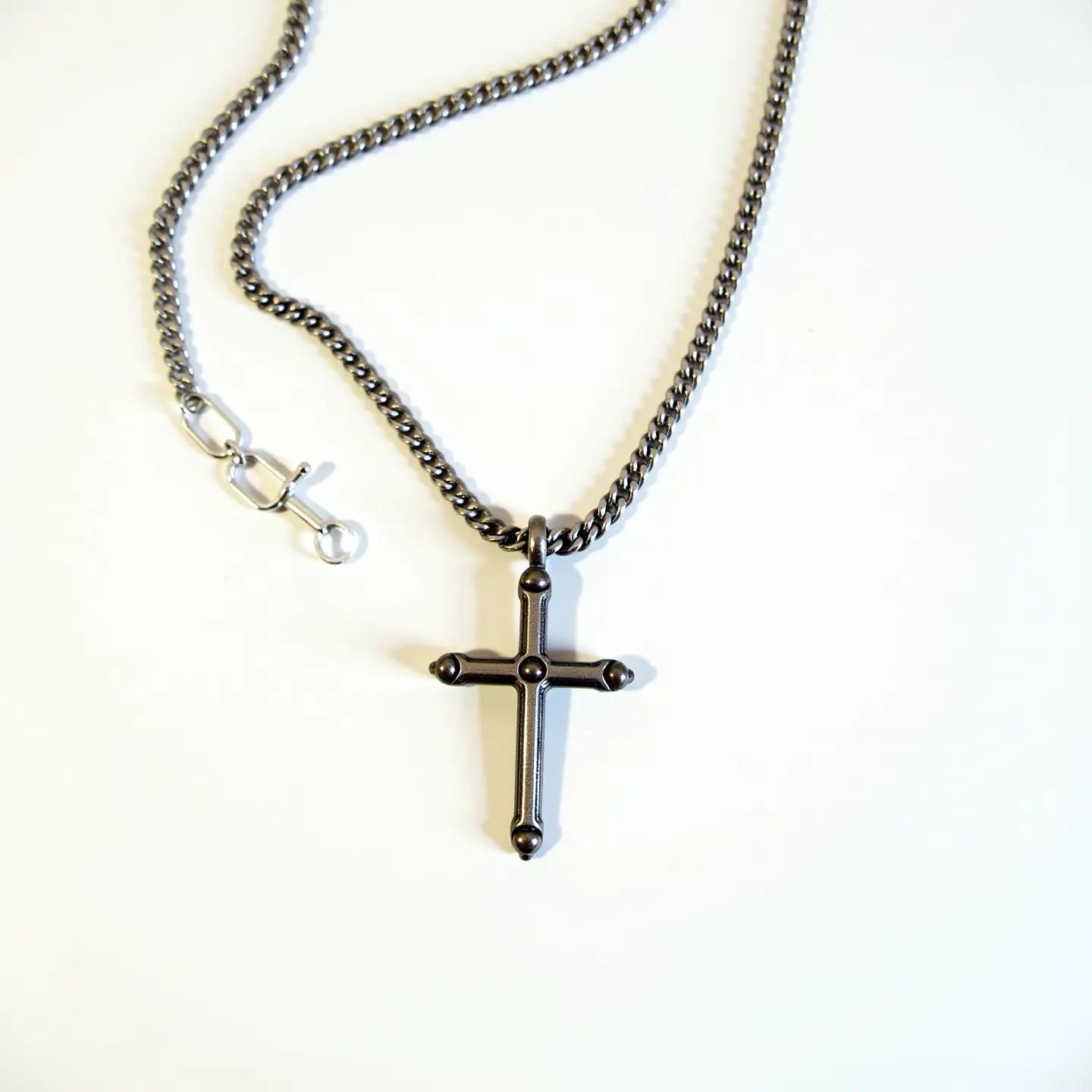 This boy's chain necklace features a robust, dark-toned metal construction, suggesting materials such as stainless steel or possibly titanium for durability and a sleek appearance. The chain is a traditional curb link style, known for its interlocking, twisted links that lay flat when worn. The necklace is adorned with a cross pendant, which mirrors the chain's metal, enhancing the cohesive design. The pendant is designed with small spherical accents at each end of the cross, adding a subtle decorative element. The necklace secures with a lobster clasp, known for its reliability and ease of use. The overall design combines practicality with a classic aesthetic.