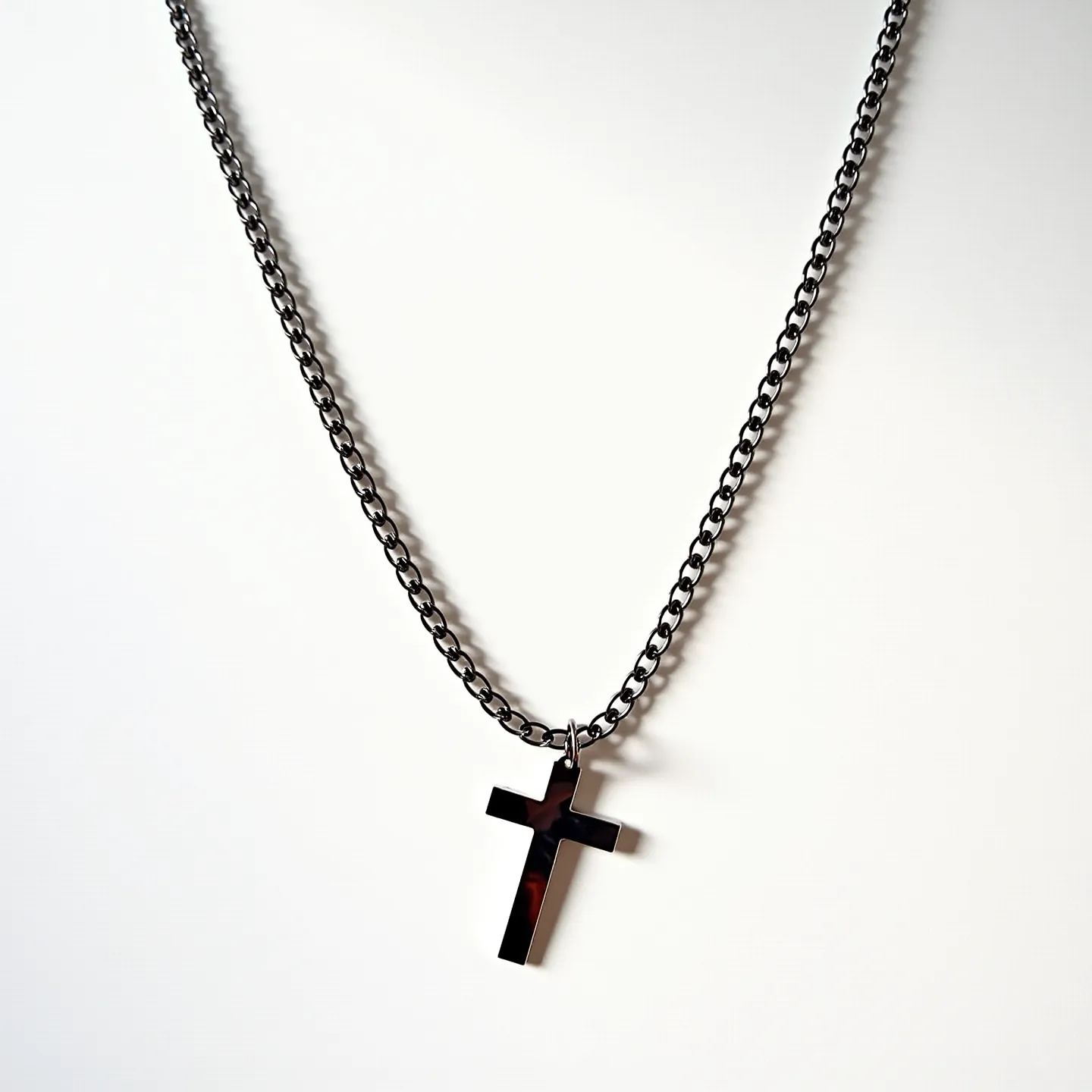 This boy's chain necklace features a dark metal chain with evenly spaced, closely linked loops, providing a robust yet stylish appearance. The necklace showcases a sleek, polished cross pendant, which hangs centrally from the chain, attached through a simple loop attachment. The cross appears to be crafted from the same metal as the chain, maintaining a cohesive and streamlined design. There are no visible gems or ornate settings present on the necklace. The overall aesthetic is modern and understated, making it a versatile accessory.