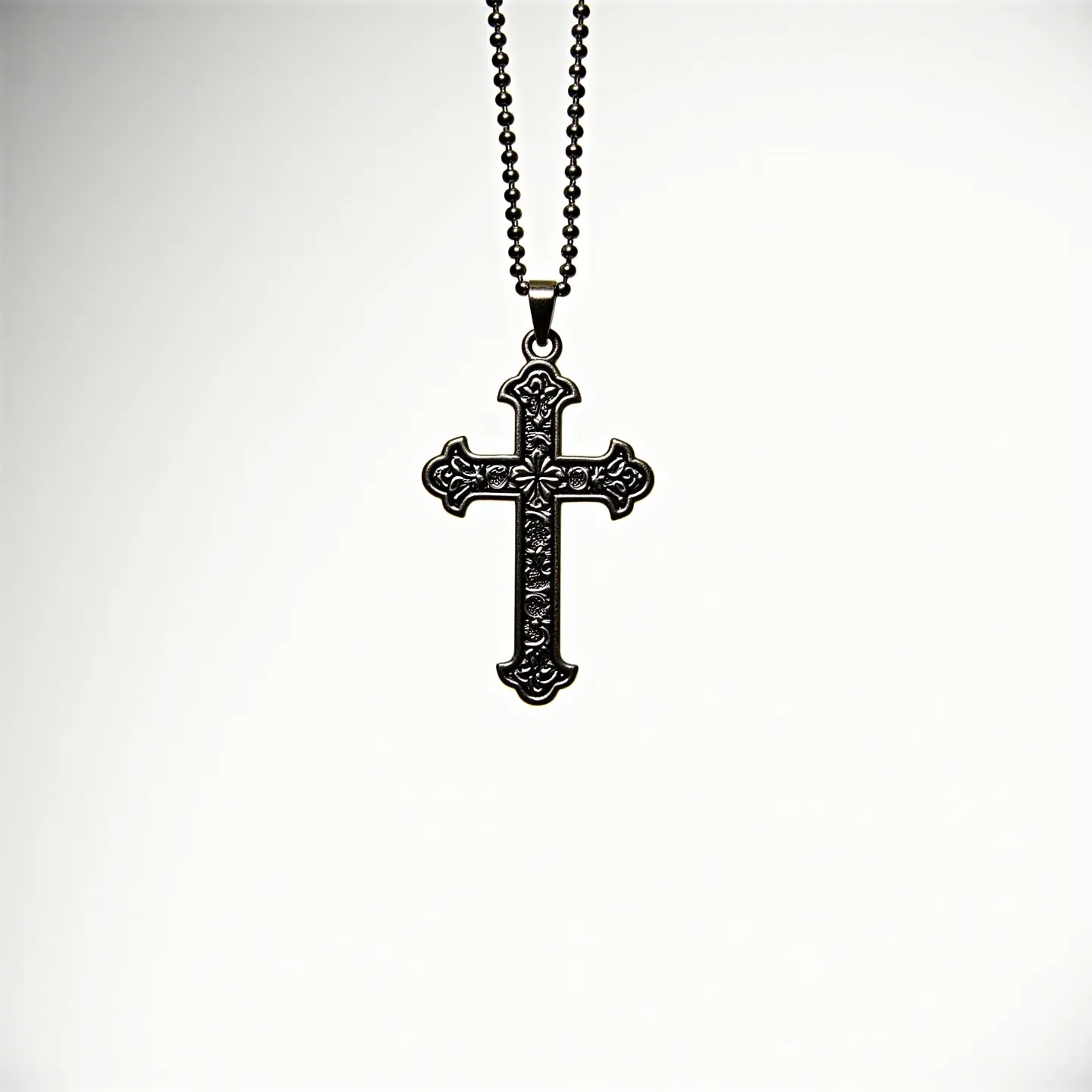 This boy's cross necklace features an intricately designed pendant with ornate detailing, crafted from a dark metallic material that gives it a robust and stylish appearance. The cross itself has a traditional shape, accented with floral motifs at each end, enhancing its decorative charm. The chain is composed of small spherical metal beads, adding a modern touch and a comfortable fit around the neck. The overall design suggests a blend of traditional and contemporary styles, making it suitable for everyday wear. The necklace is finished with a simple metal loop at the top of the cross for secure attachment to the chain.