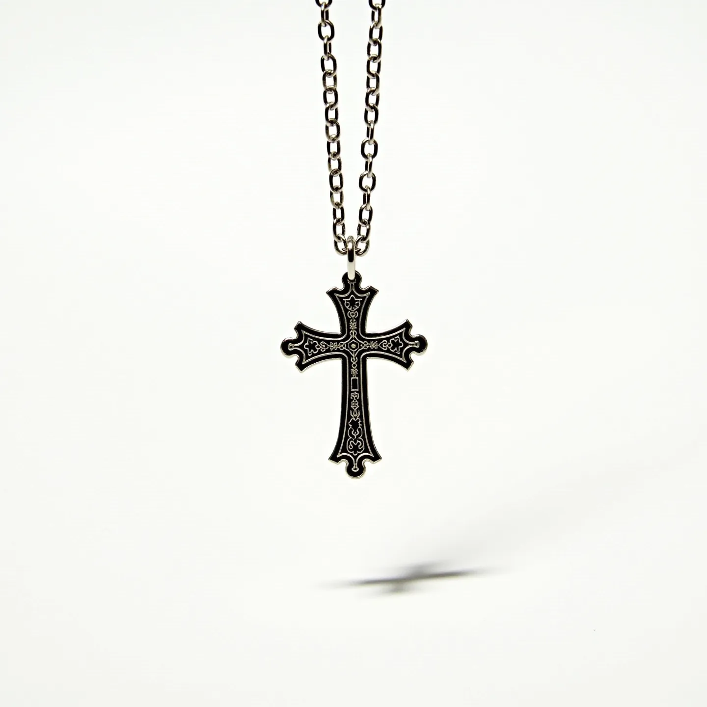 This boy's cross necklace features an intricately designed cross pendant, crafted from a metal with a dark finish that highlights its intricate engravings. The cross does not appear to include any gemstones, as its main decorative elements are the detailed patterns etched into the metal itself. The pendant hangs from a sturdy chain, also made of metal, composed of uniform, oval-shaped links. The connection between the chain and the cross is achieved through a simple loop at the top of the pendant, allowing for smooth movement and wearability. The necklace's overall craftsmanship suggests durability and attention to decorative detail.