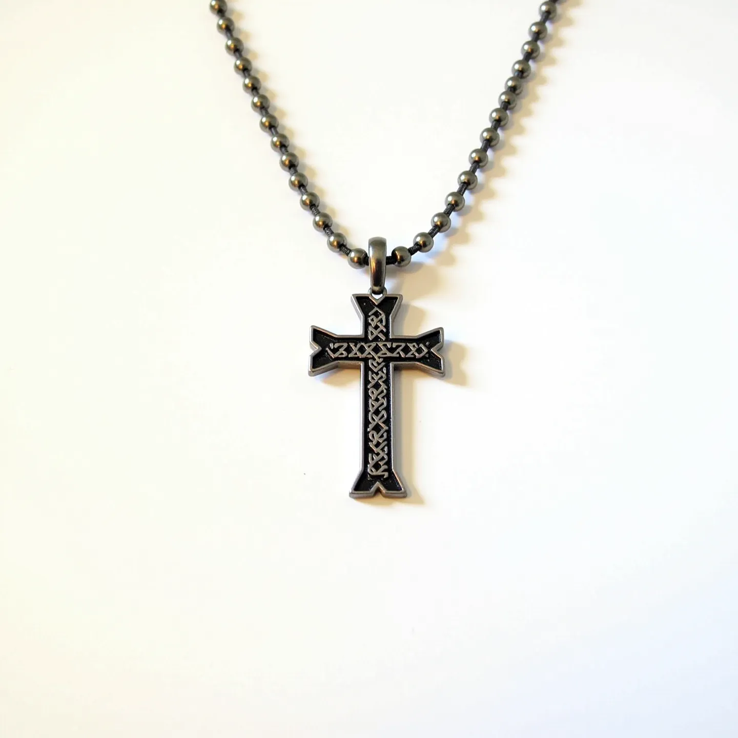 This boy's cross necklace features a black metallic chain with a classic cross pendant. The pendant is designed with intricate interwoven patterns along its surface, adding a detailed and sophisticated aesthetic. The material of the pendant appears to be metal with a dark finish, likely stainless steel or a similar durable alloy, enhancing its longevity and resistance to tarnish. The chain consists of uniformly rounded beads or links, contributing to a robust and masculine appearance. The necklace is equipped with a simple loop attachment that connects the pendant to the chain, ensuring that it remains securely fastened during wear.