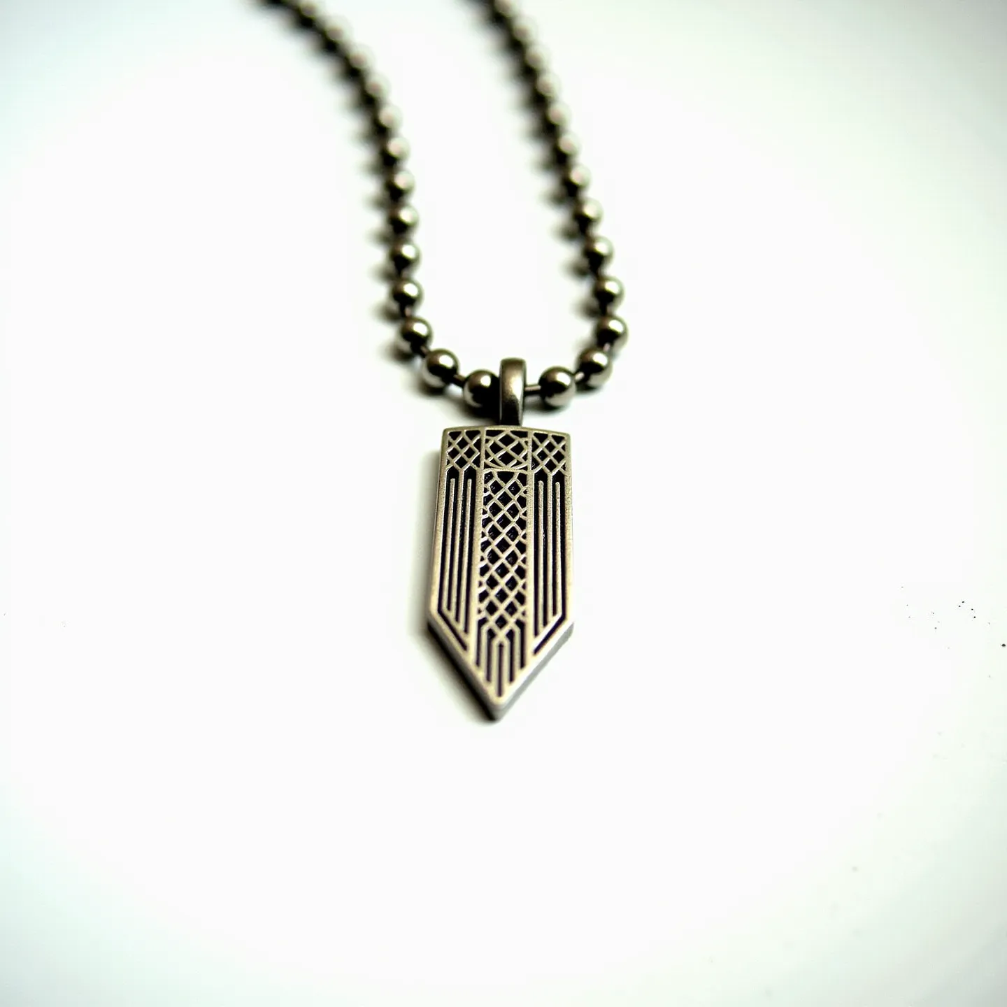 This boy's necklace features a pendant in a shield-like shape with intricate geometric patterns etched into its surface, giving it a modern and edgy appearance. The pendant, likely made of metal due to its sheen and structure, hangs from a beaded chain. The beads appear metallic, matching the pendant in color and style, suggesting a unified design. The pendant is looped onto the chain through a small, sturdy-looking bail. The necklace overall conveys a bold and contemporary look, suitable for casual or statement wear.