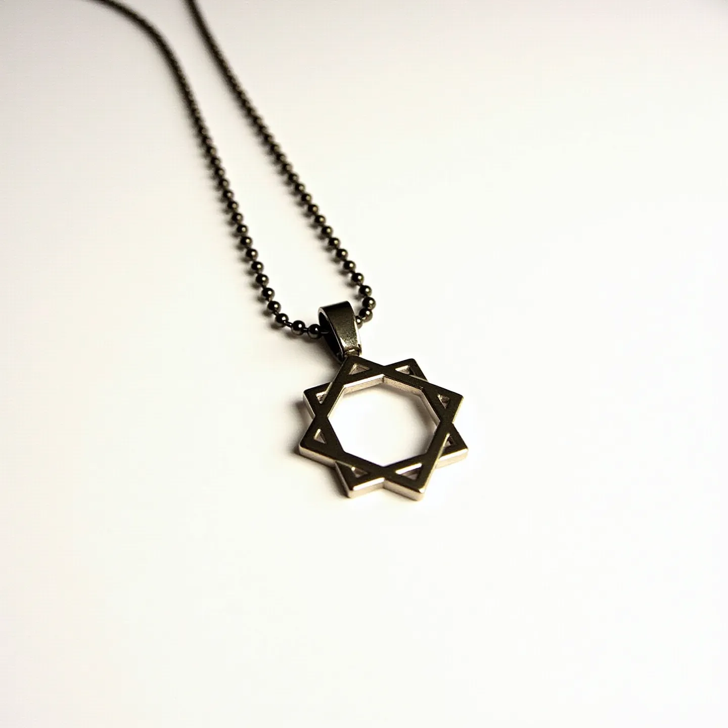 This boy's necklace features a distinctive pendant crafted from a metallic material that forms a geometric star design, adding a modern touch to the piece. The pendant is suspended from a ball chain, also made of a similar metal, which gives the necklace a cohesive look. The star pendant is open and minimalist, without any gems or stones. The chain is likely equipped with a simple clasp or connector, although it is not prominently visible in the image. The overall design has a sleek and contemporary aesthetic.